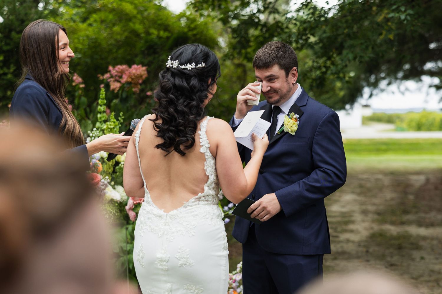 Emotional groom wipes tears during outdoor wedding vows while officiant and bride look on warmly.