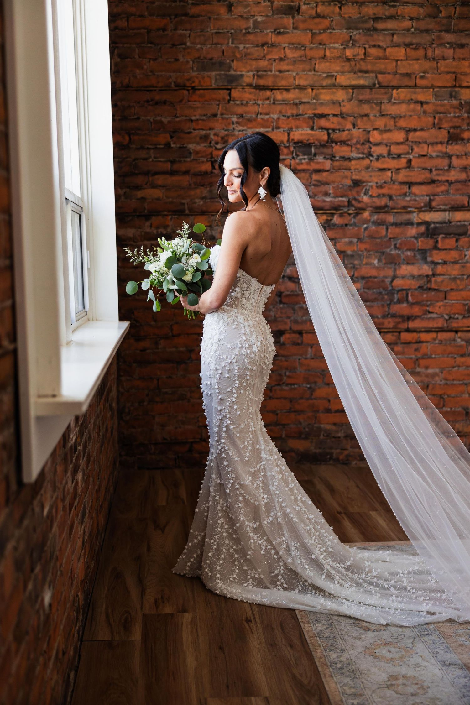 Elegant bridal portrait against exposed brick wall featuring detailed lace gown and flowing veil.