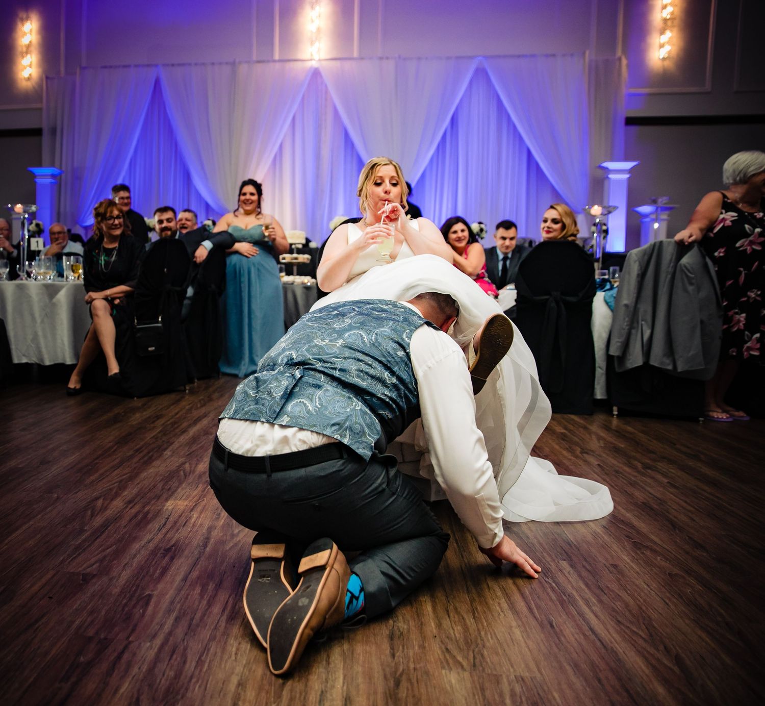 Traditional wedding garter removal moment during reception on wooden dance floor with blue backdrop.