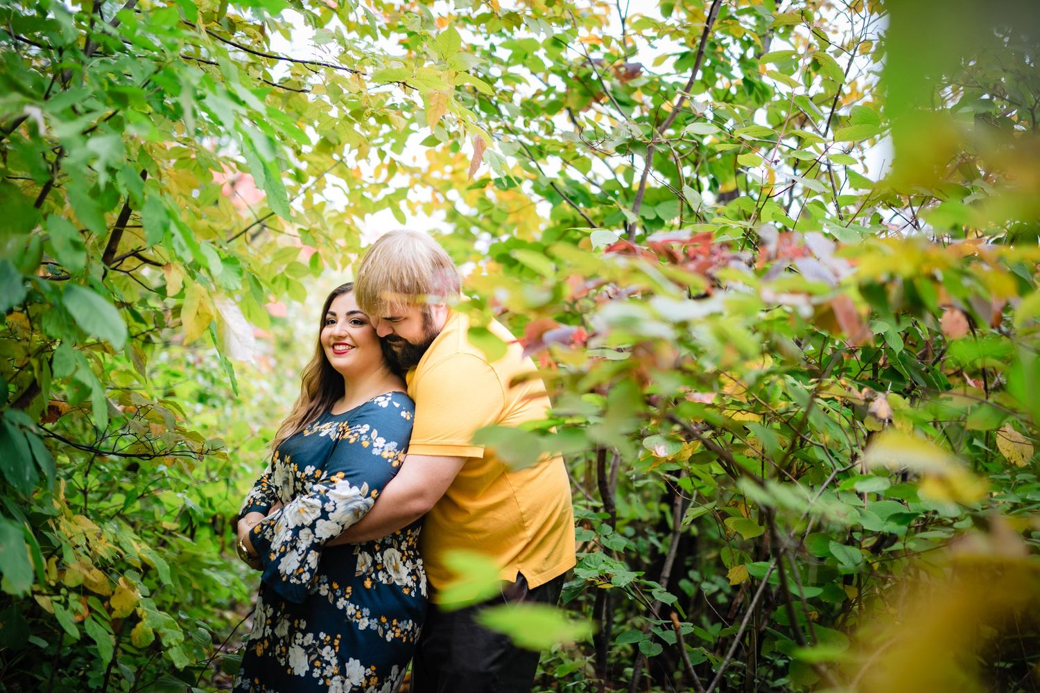 Romantic engagement photos captured through green foliage in golden sunlight.