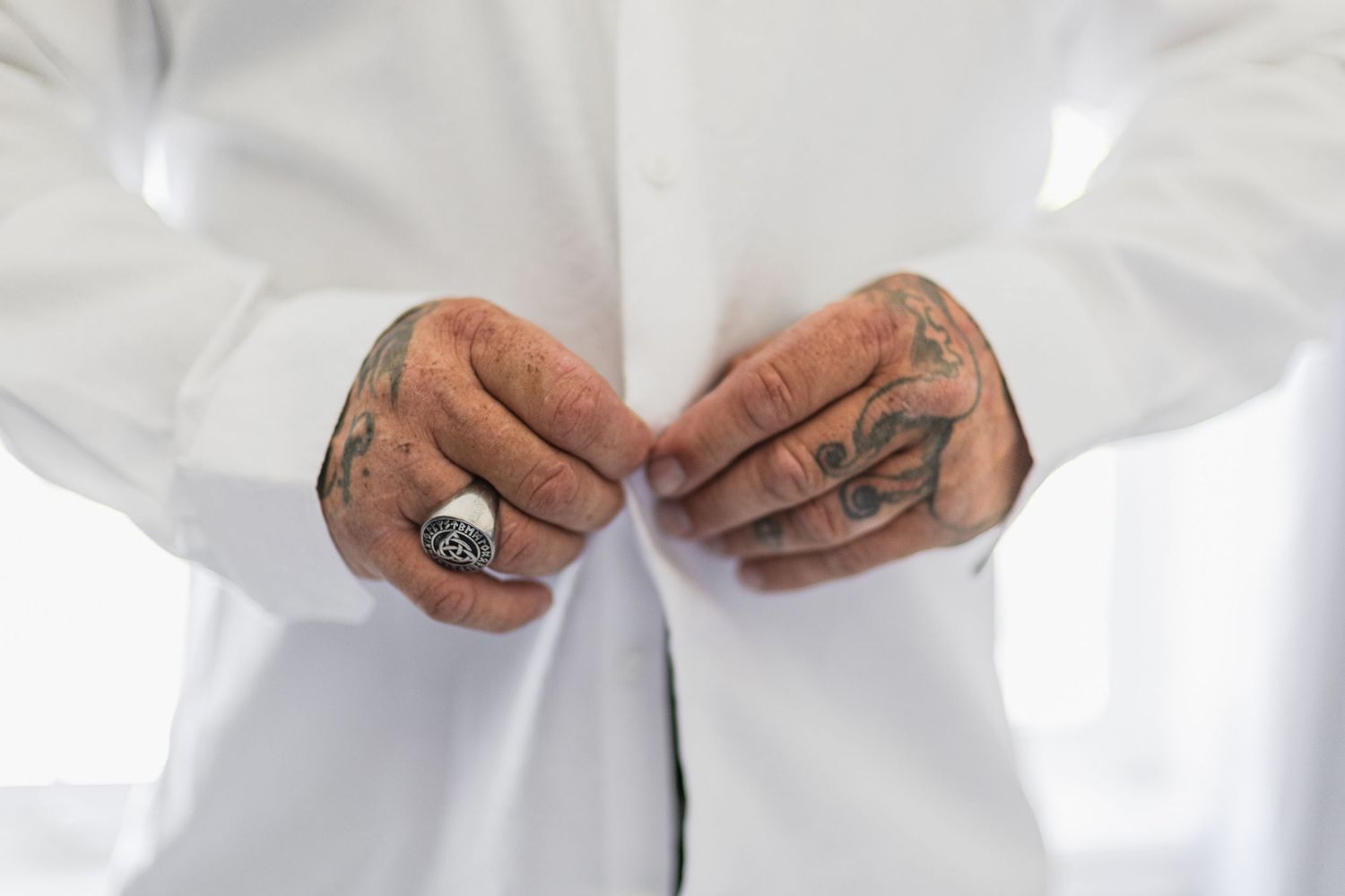 Groom with tattooed hands fastening white dress shirt buttons before wedding ceremony.
