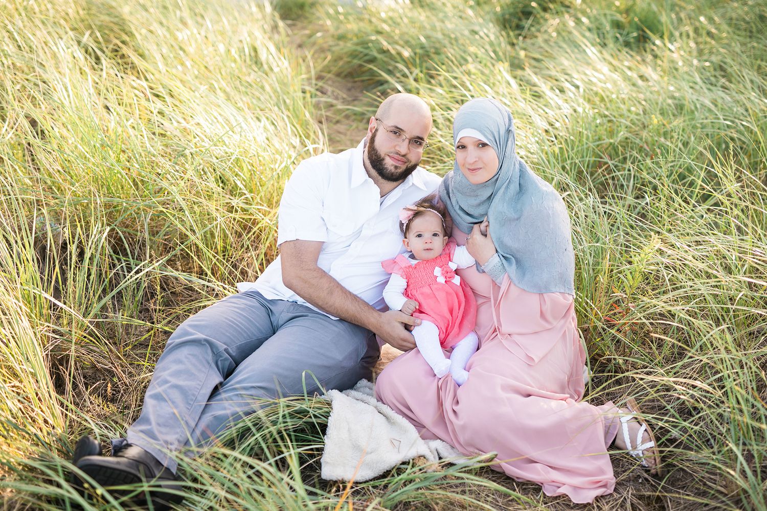 Family poses together in tall grass field wearing coordinated pink and neutral outfits.