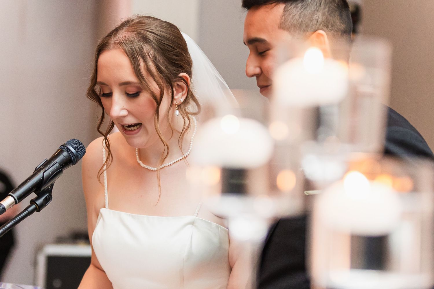 Bride in white dress and veil speaking at microphone during wedding reception with candles in foreground.