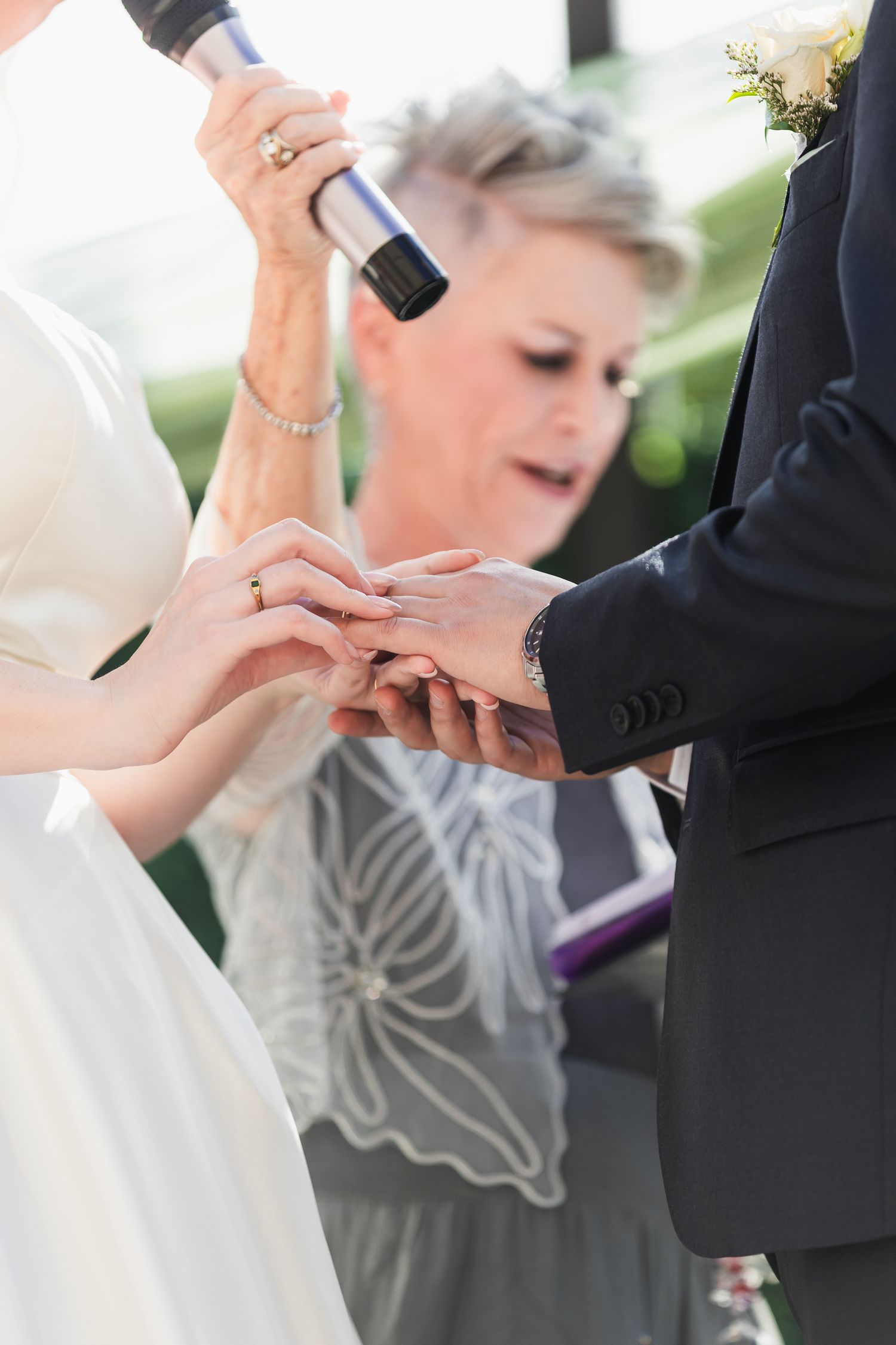 Officiant places wedding ring on bride's finger during outdoor ceremony while holding microphone.