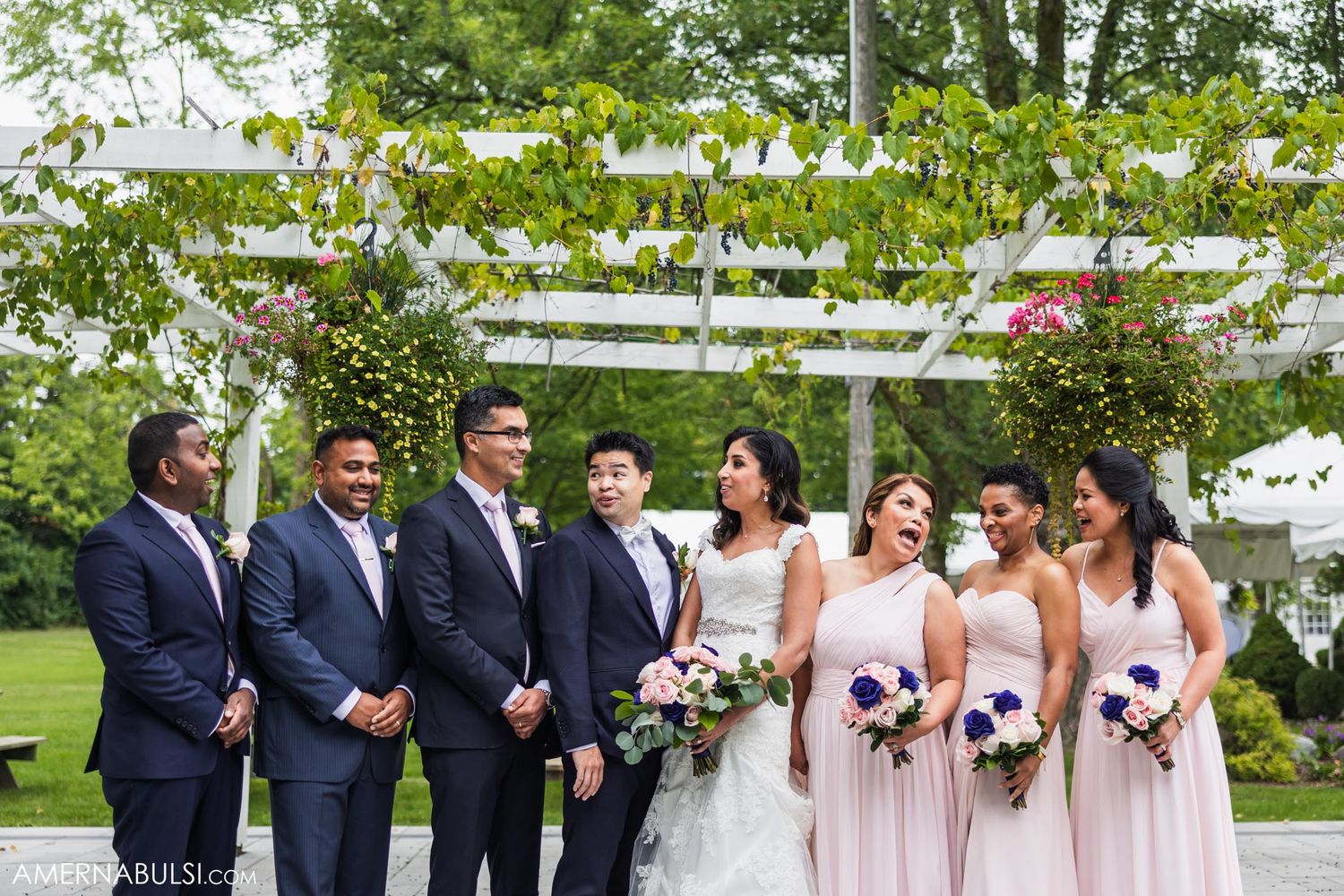 Full wedding party in navy and pink attire poses outdoors under white pergola.