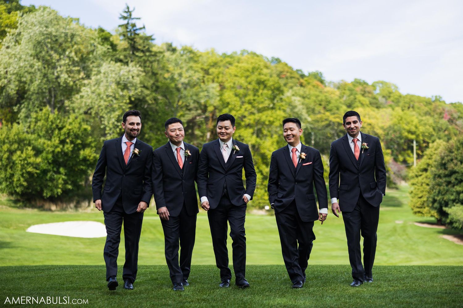 Groomsmen in matching navy suits walk together on grass with trees in background.