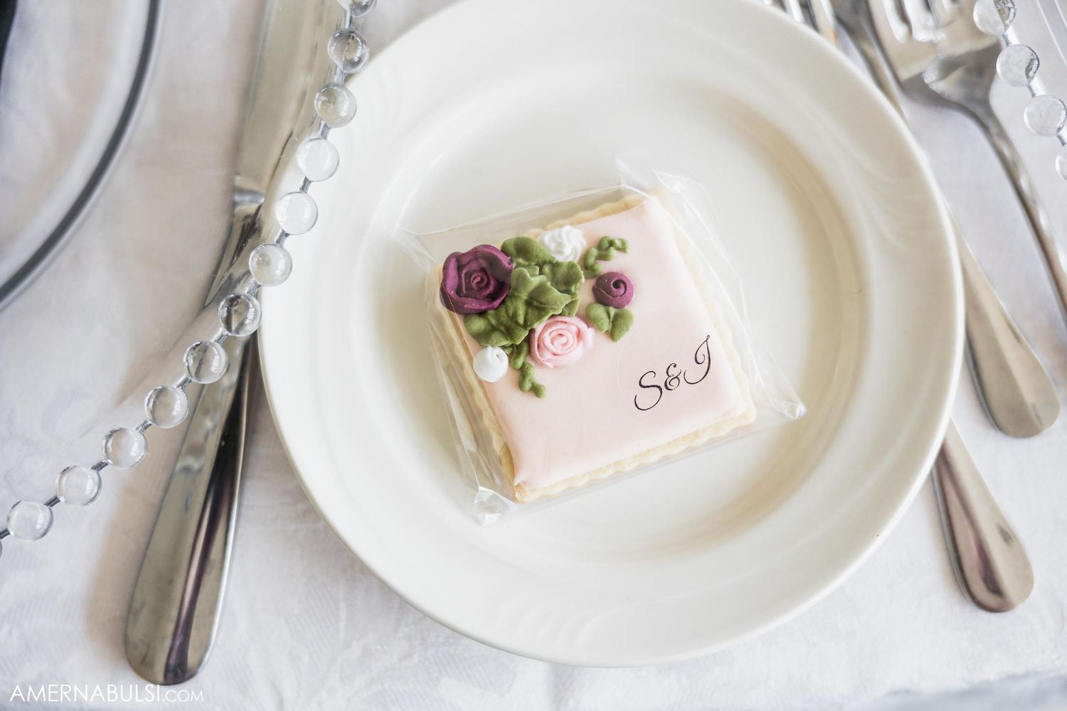 A pink and white decorated mini cake or petit four on an elegant white plate with silverware.