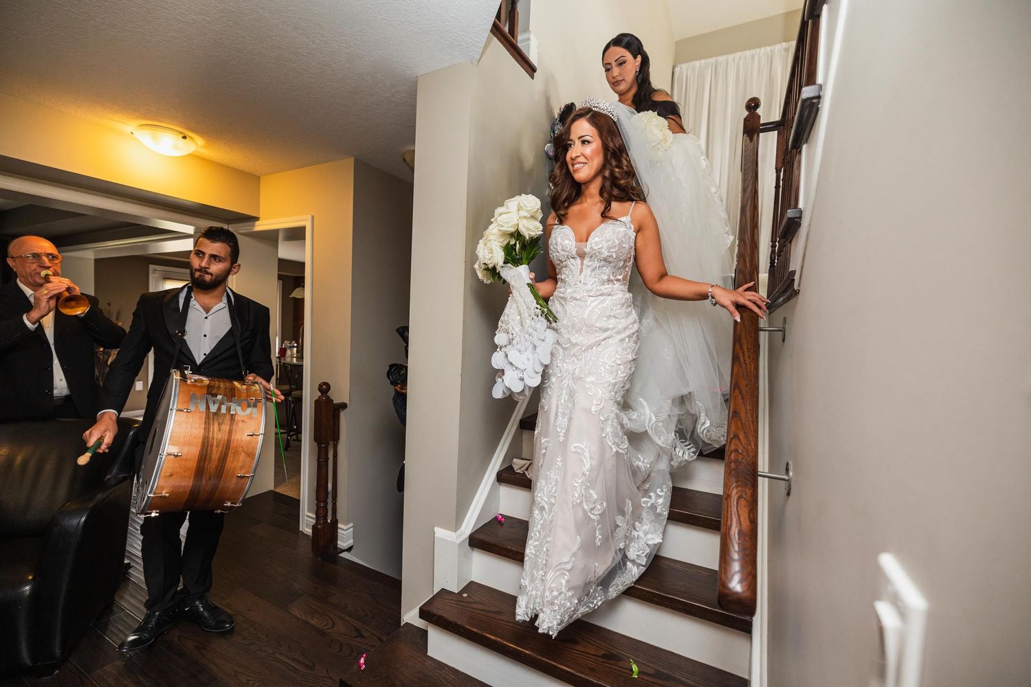 Person in a lace wedding gown descends wooden stairs while holding white flowers.
