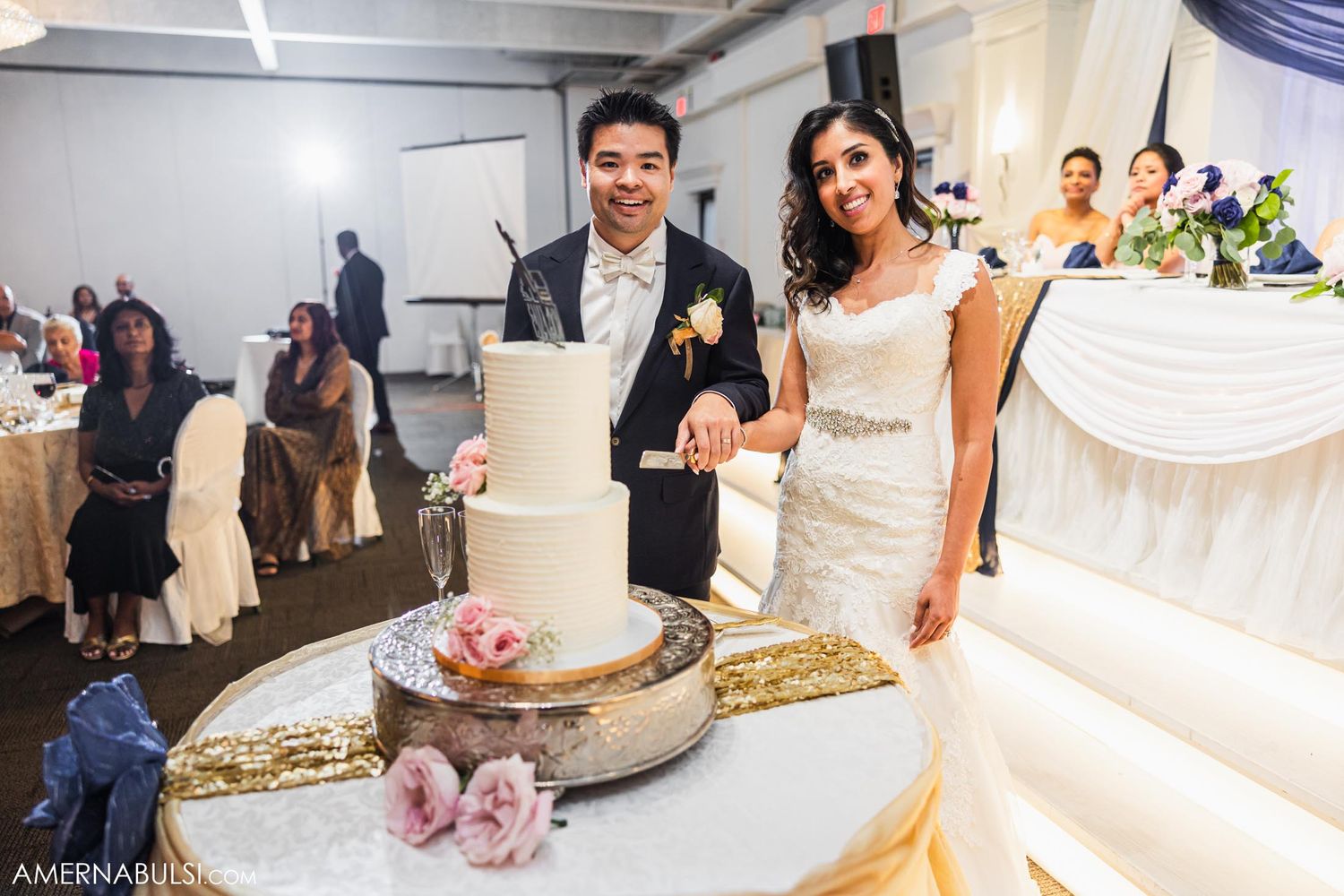 Happy couple cutting their three-tiered white wedding cake decorated with pink roses.