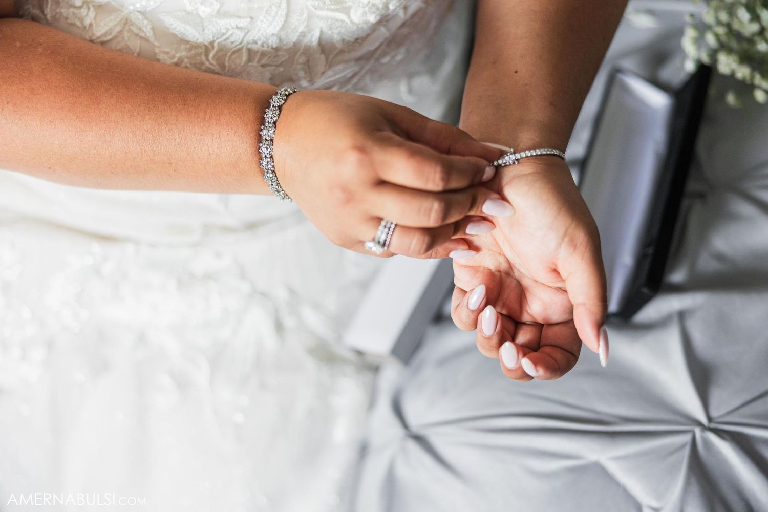 Close-up of hands wearing an engagement ring and bracelet against white fabric.
