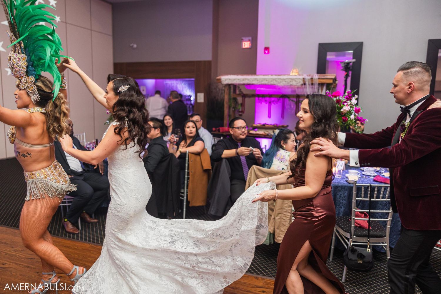 Wedding party dancing energetically on dance floor with showgirl performer in festive mood with purple lighting.