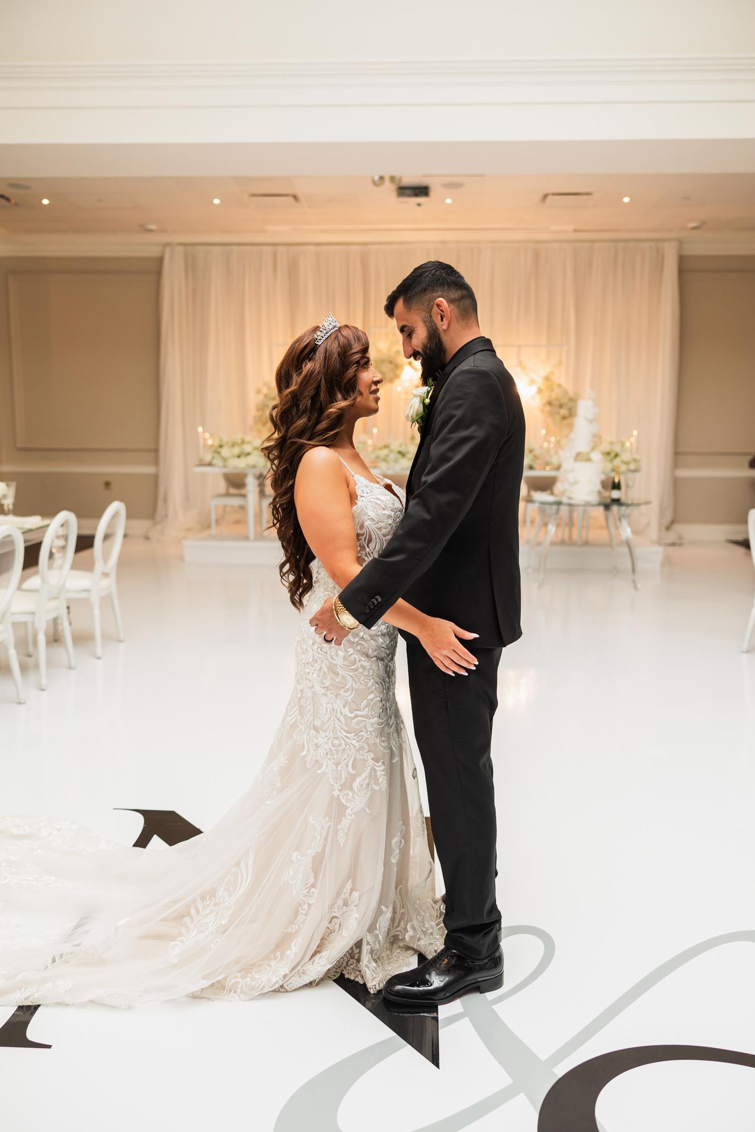 A couple in wedding attire share an intimate moment in a white decorated ballroom.