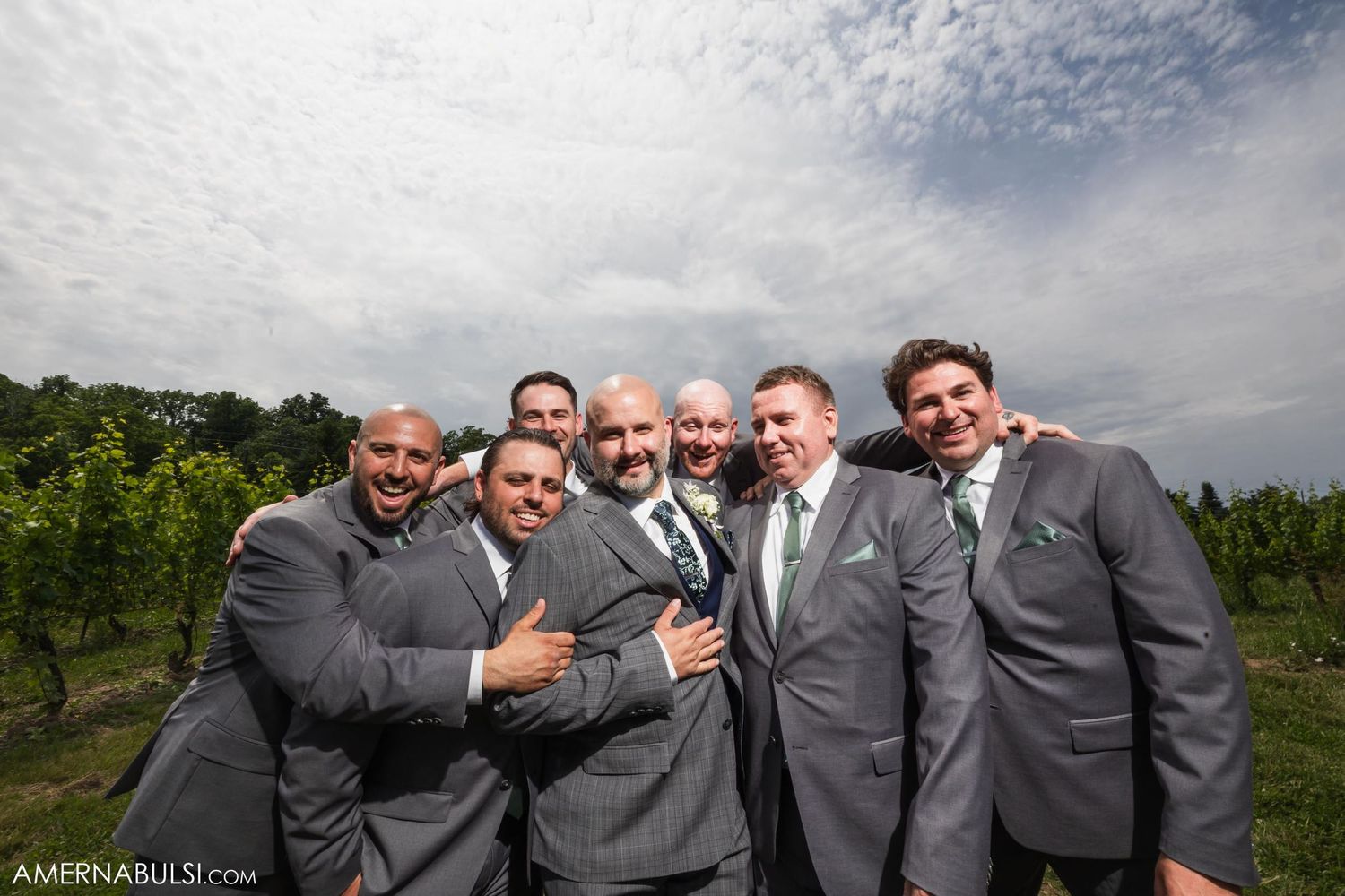 Wedding party of groomsmen in grey suits sharing a laugh in front of vineyard rows.