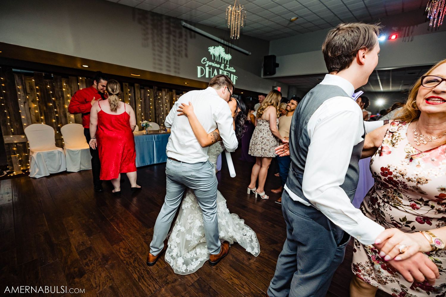 Couples dancing on wooden floor at reception with string lights in background.