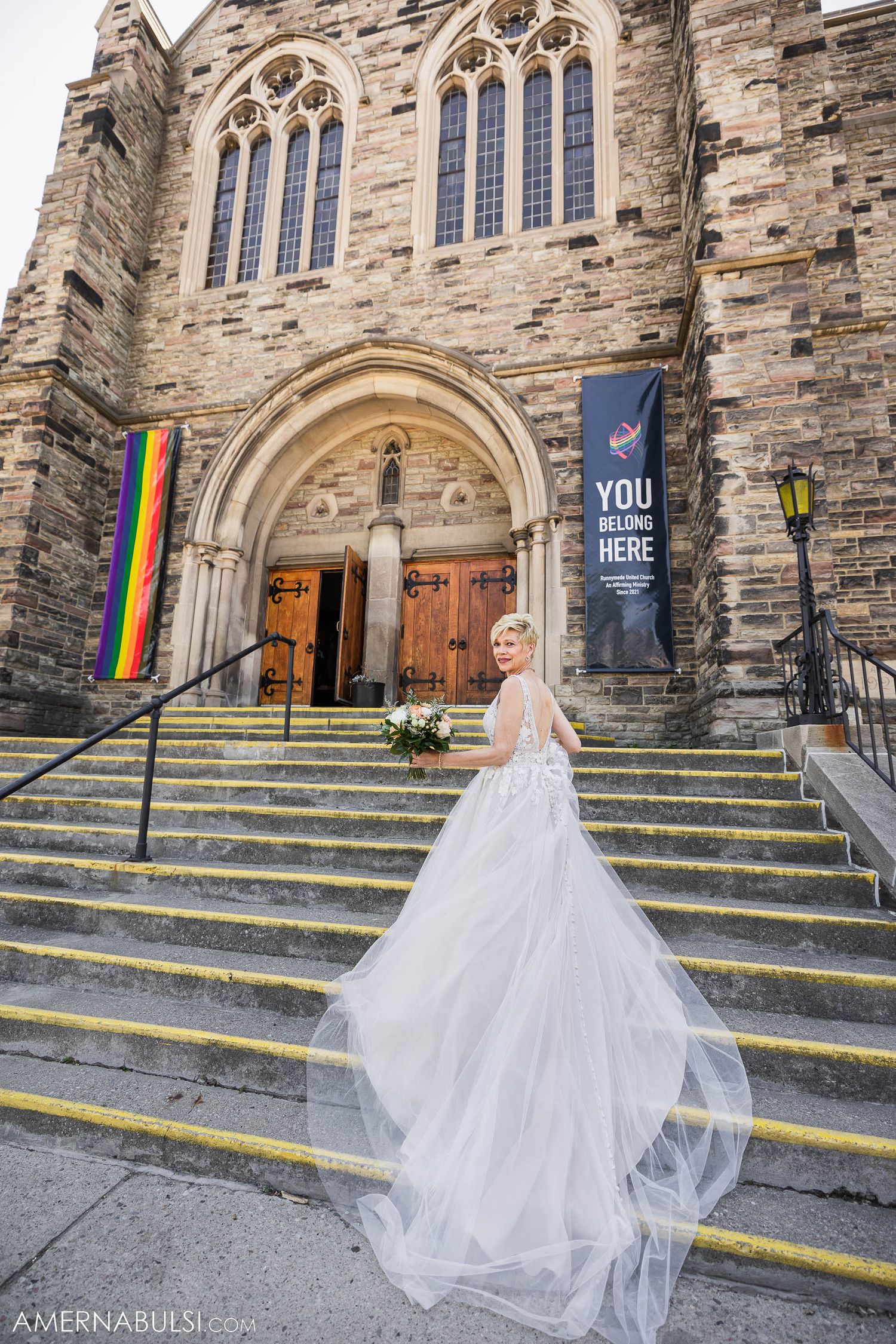 Bride in flowing dress poses on stone church steps with rainbow flag visible.