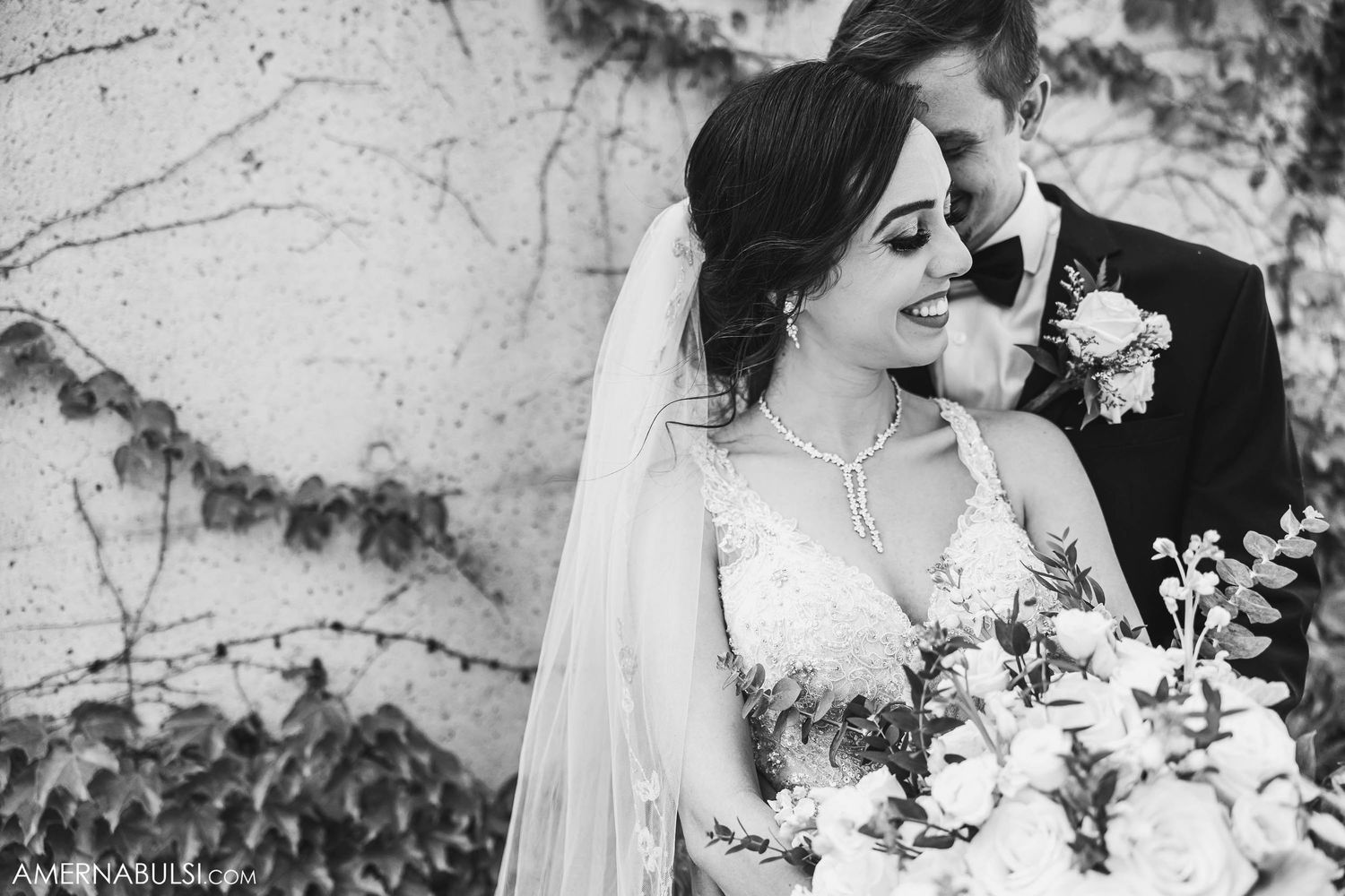Black and white portrait of a couple against an ivy-covered wall on their wedding day.