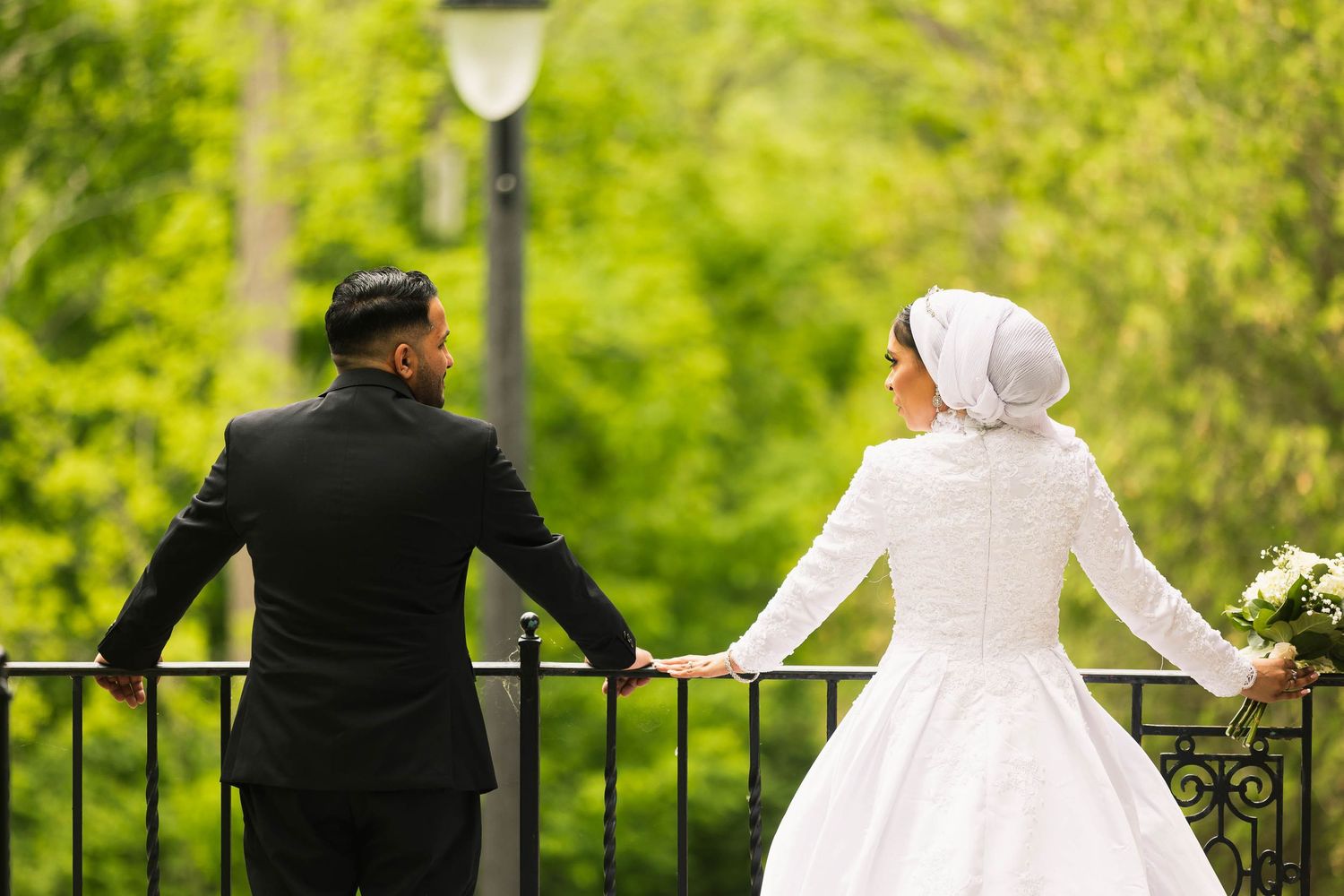 Wedding couple stands at iron railing overlooking garden with lamp post.