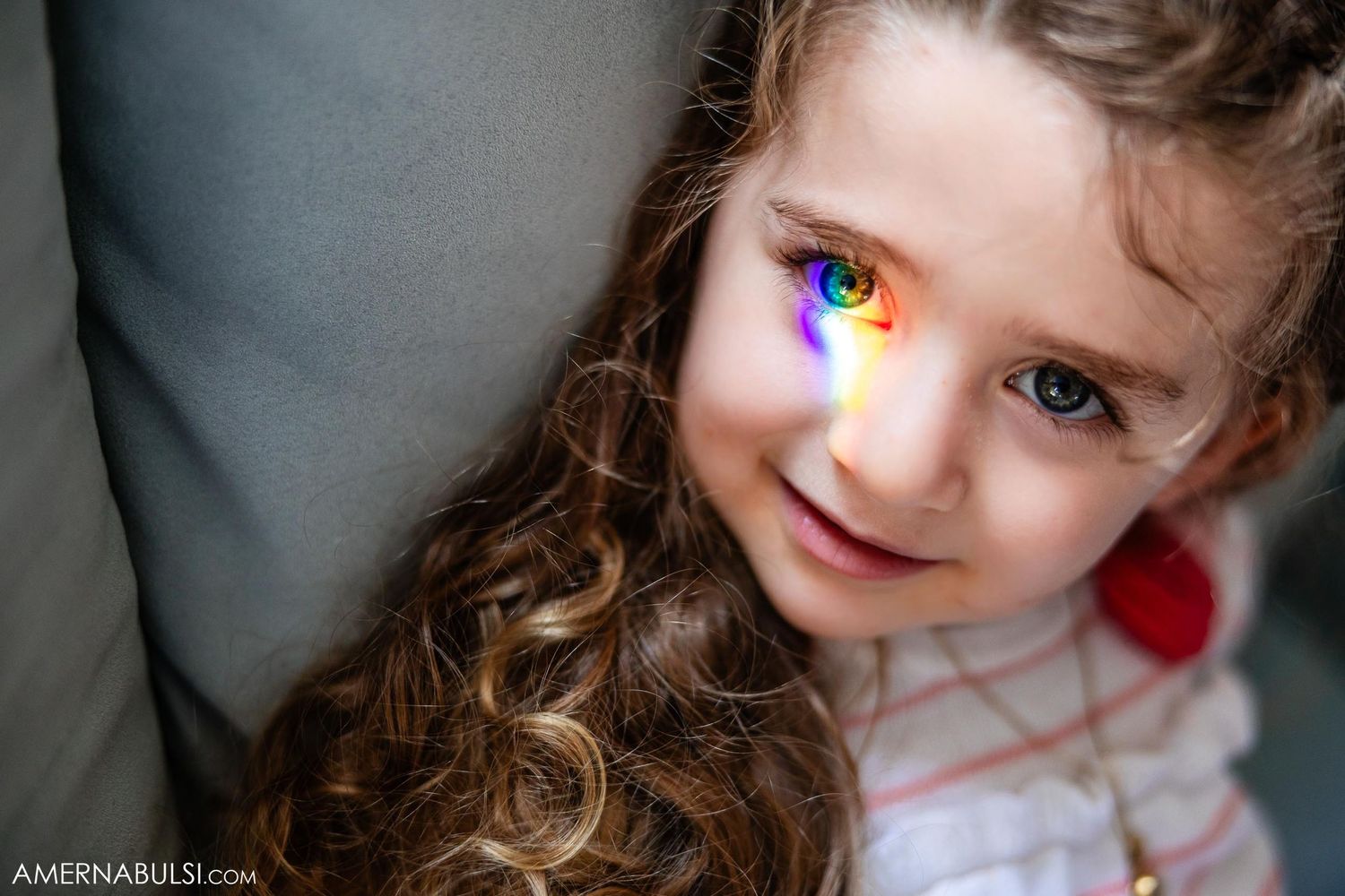 A close-up portrait of a child with long hair and a colorful rainbow reflection across their eye.