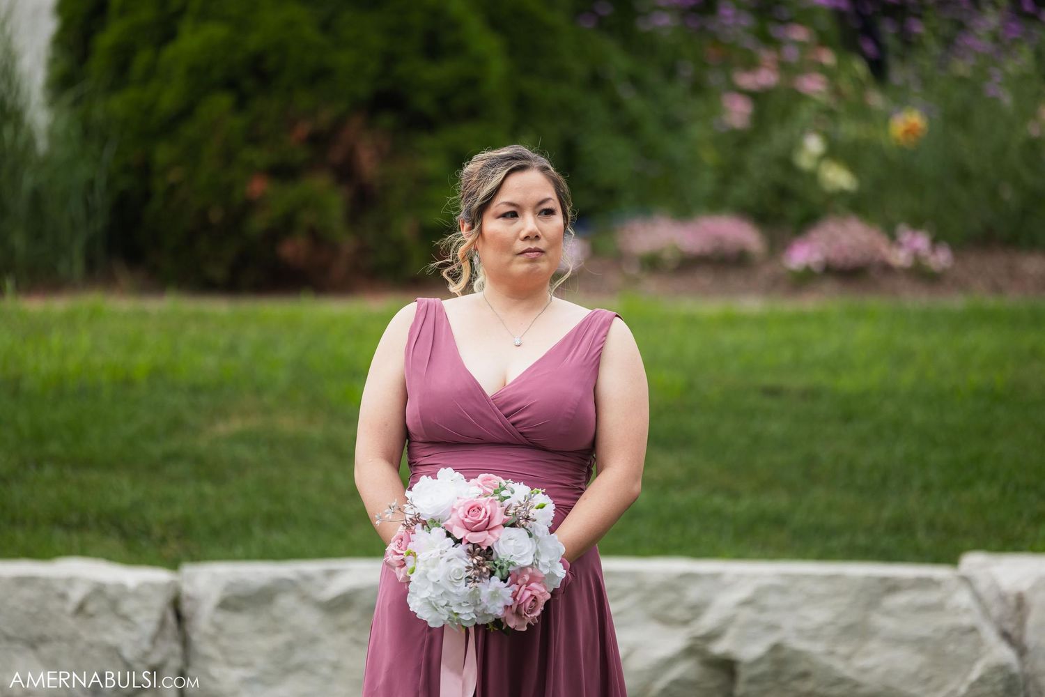 Bridesmaid in dusty rose dress holding pastel floral bouquet stands on lawn with flower garden backdrop.