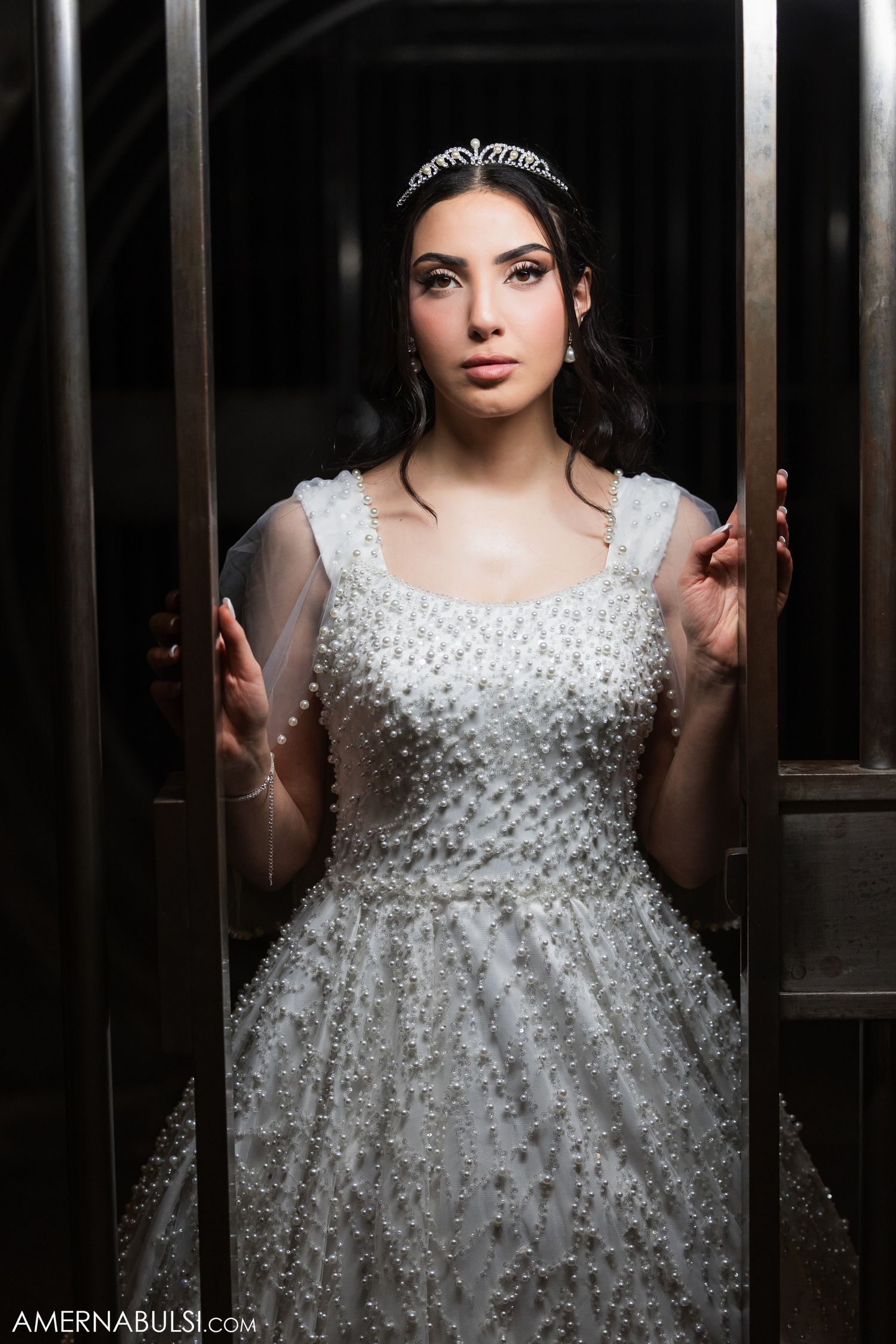 A bride in a sparkling white wedding gown with beaded details poses dramatically in a dark doorway.