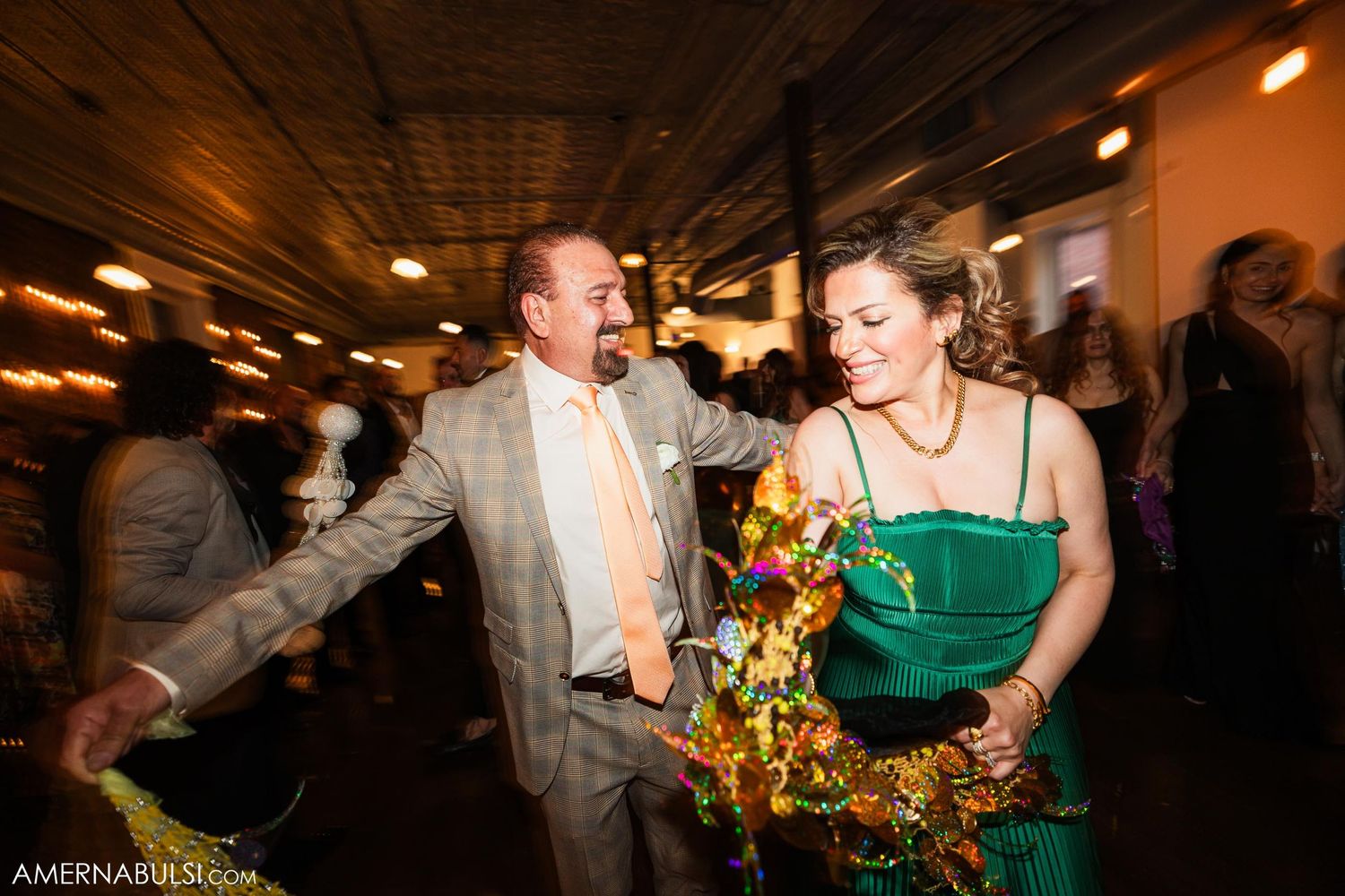Guests in formal attire dancing with festive gold decorations at an indoor wedding celebration.