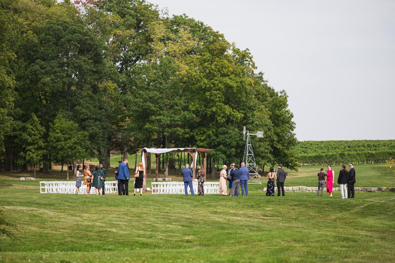 Wedding ceremony setup outdoors with guests gathering on green lawn near wooded area.