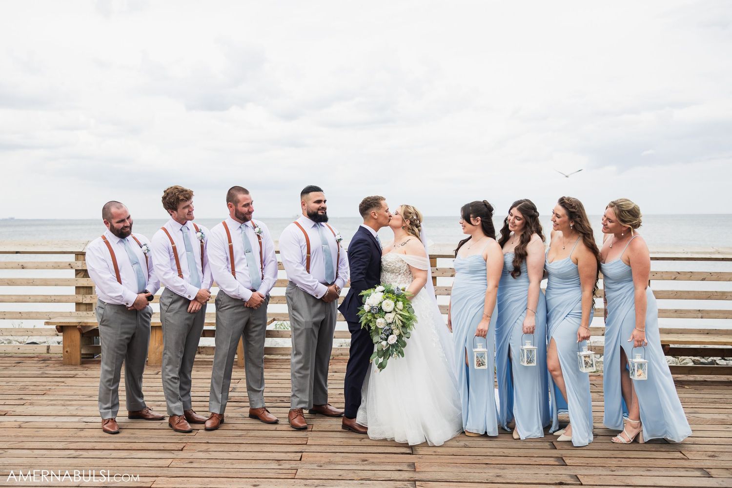 Wedding party dressed in light blue and white poses on a beachfront boardwalk.