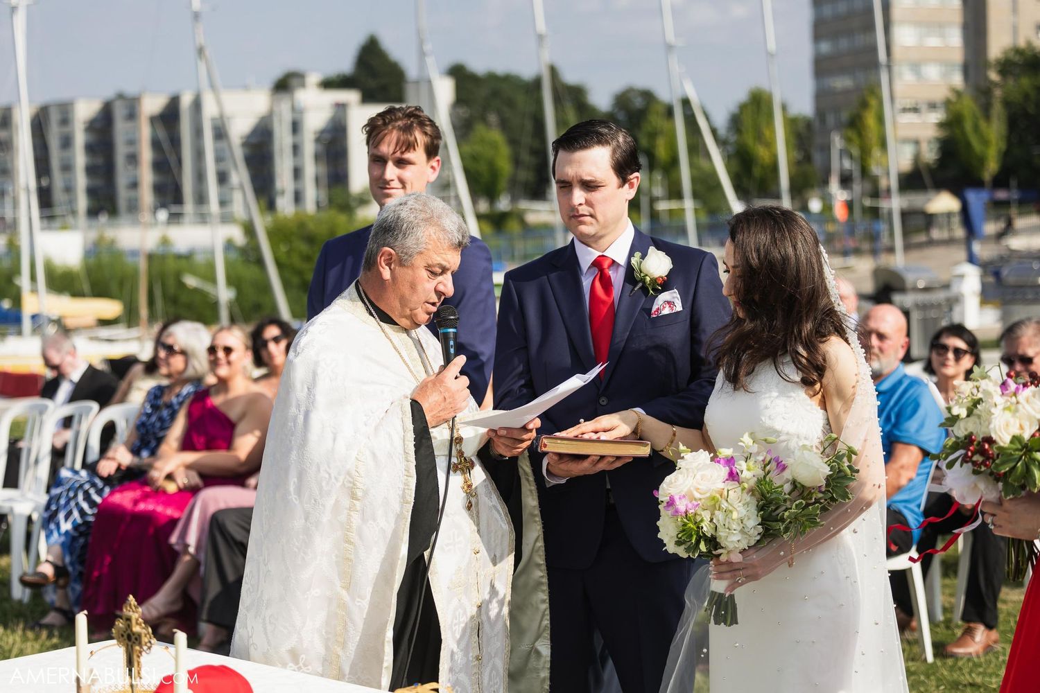 An outdoor wedding ceremony takes place by a marina with boats and buildings in the background.