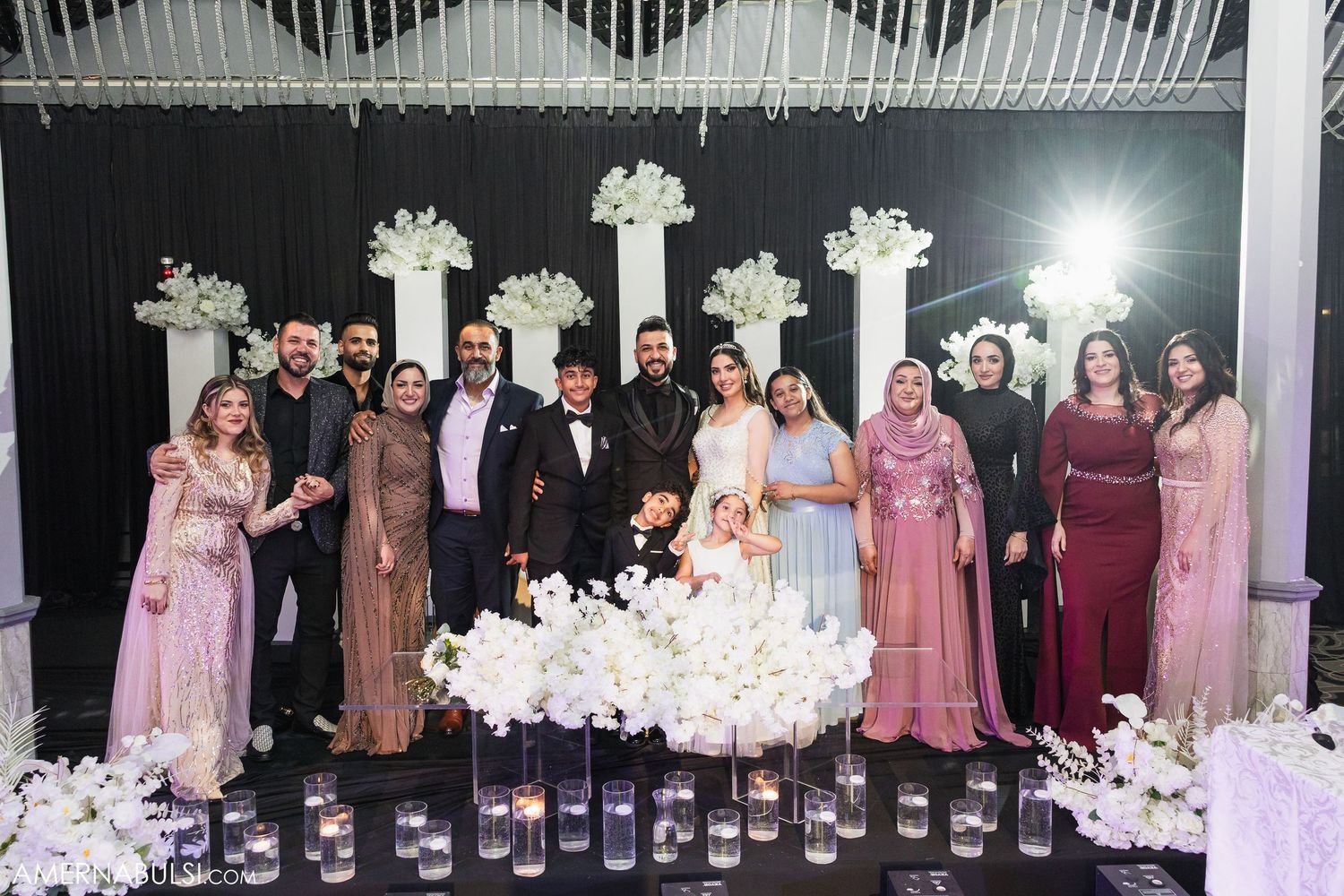 A panoramic view of wedding guests standing in a reception venue decorated with white floral arrangements and columns.