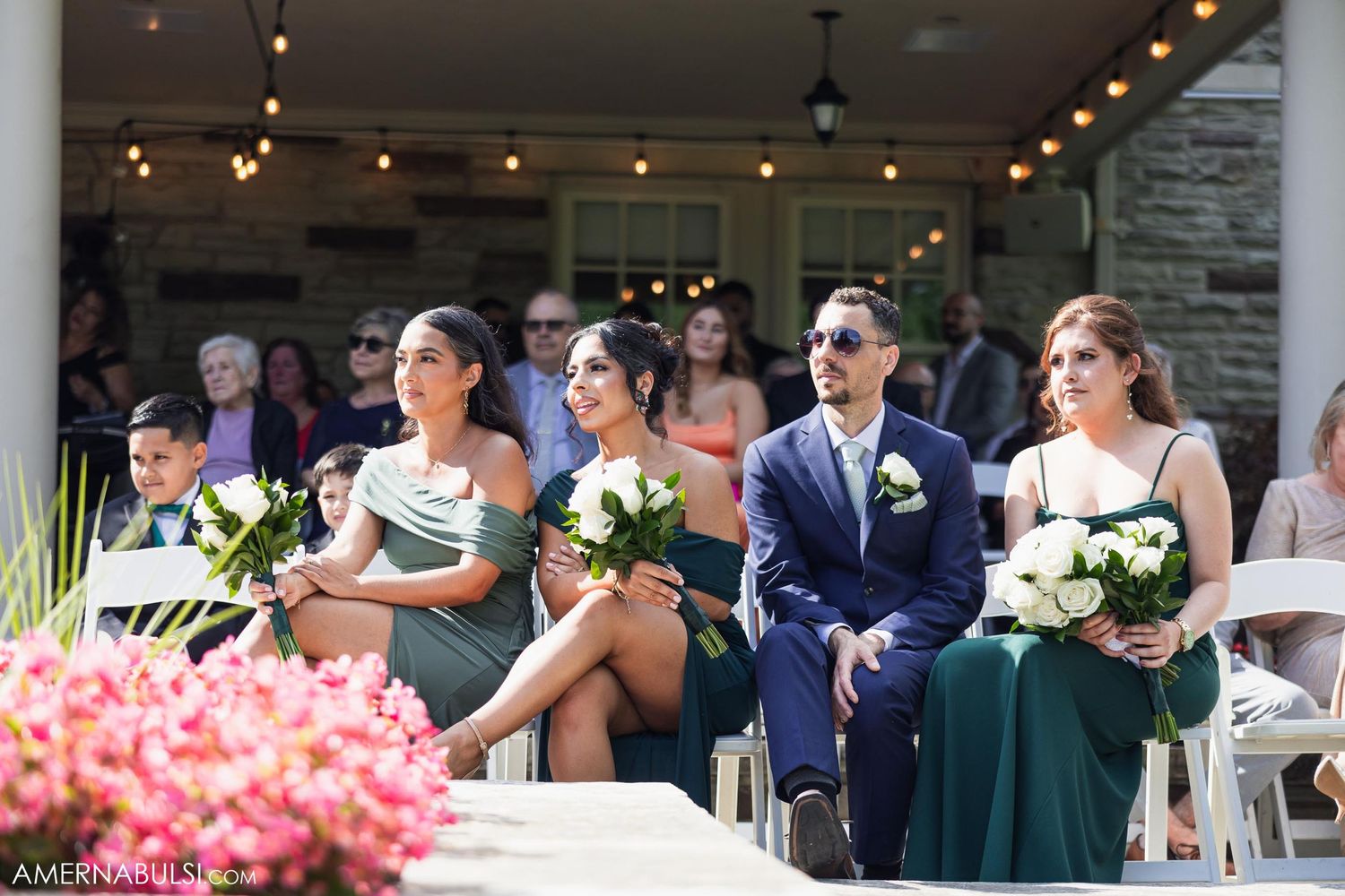 Wedding guests in formal attire seated at ceremony with floral decorations and string lights.