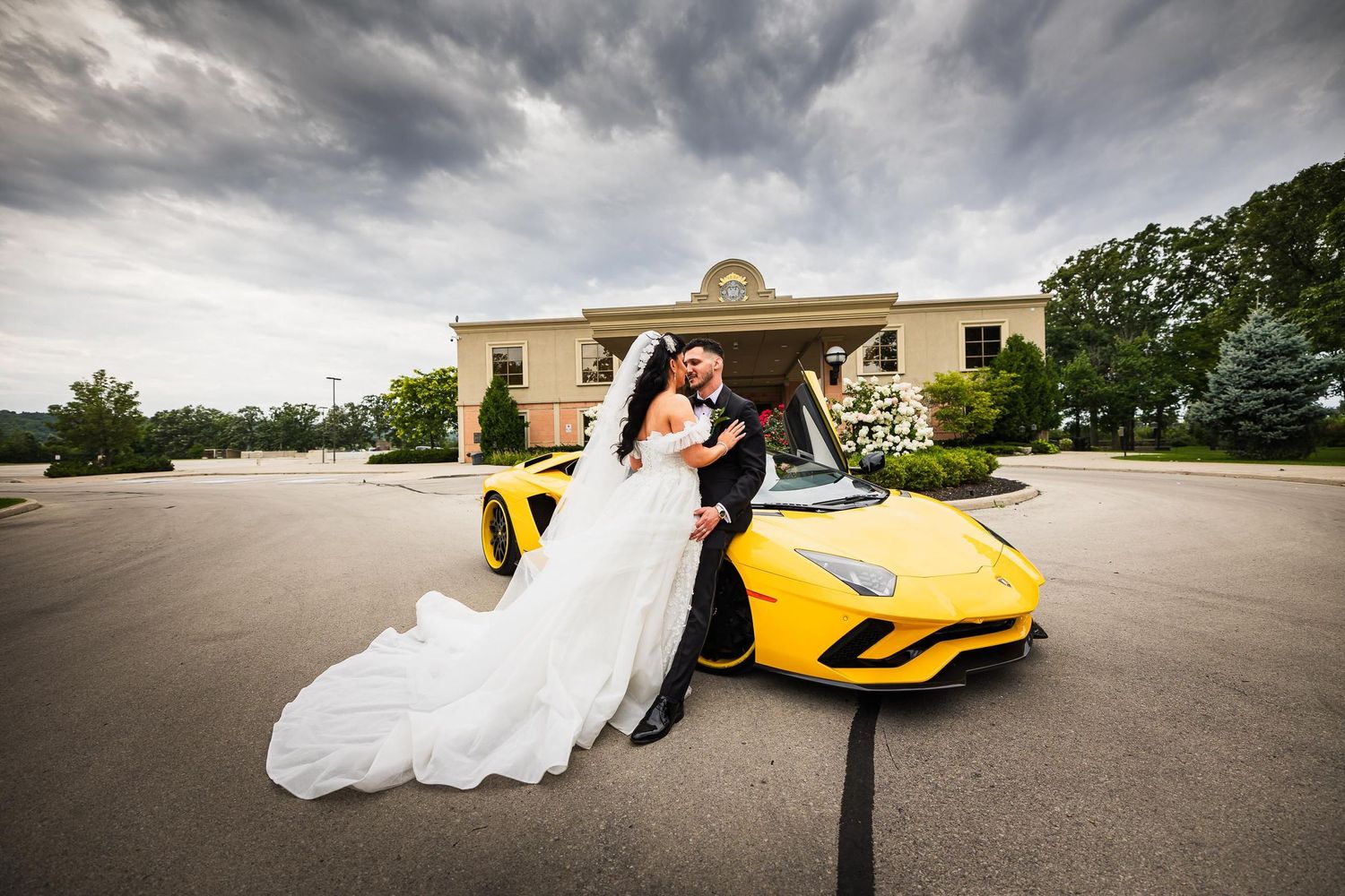 Wedding couple poses with yellow Lamborghini in parking lot under dramatic clouds.