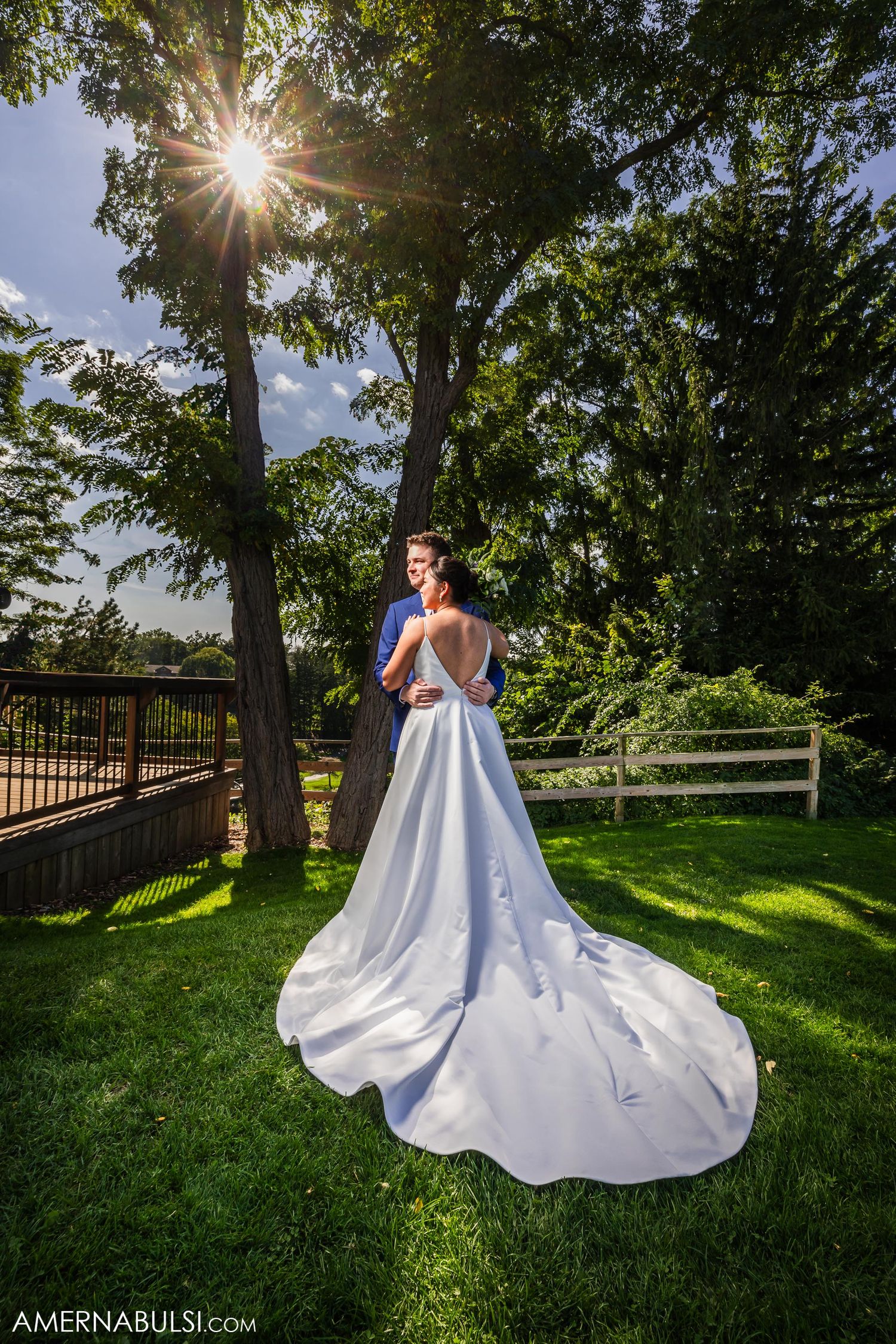 Dramatic wedding dress silhouette against sunburst through trees in lush garden setting.
