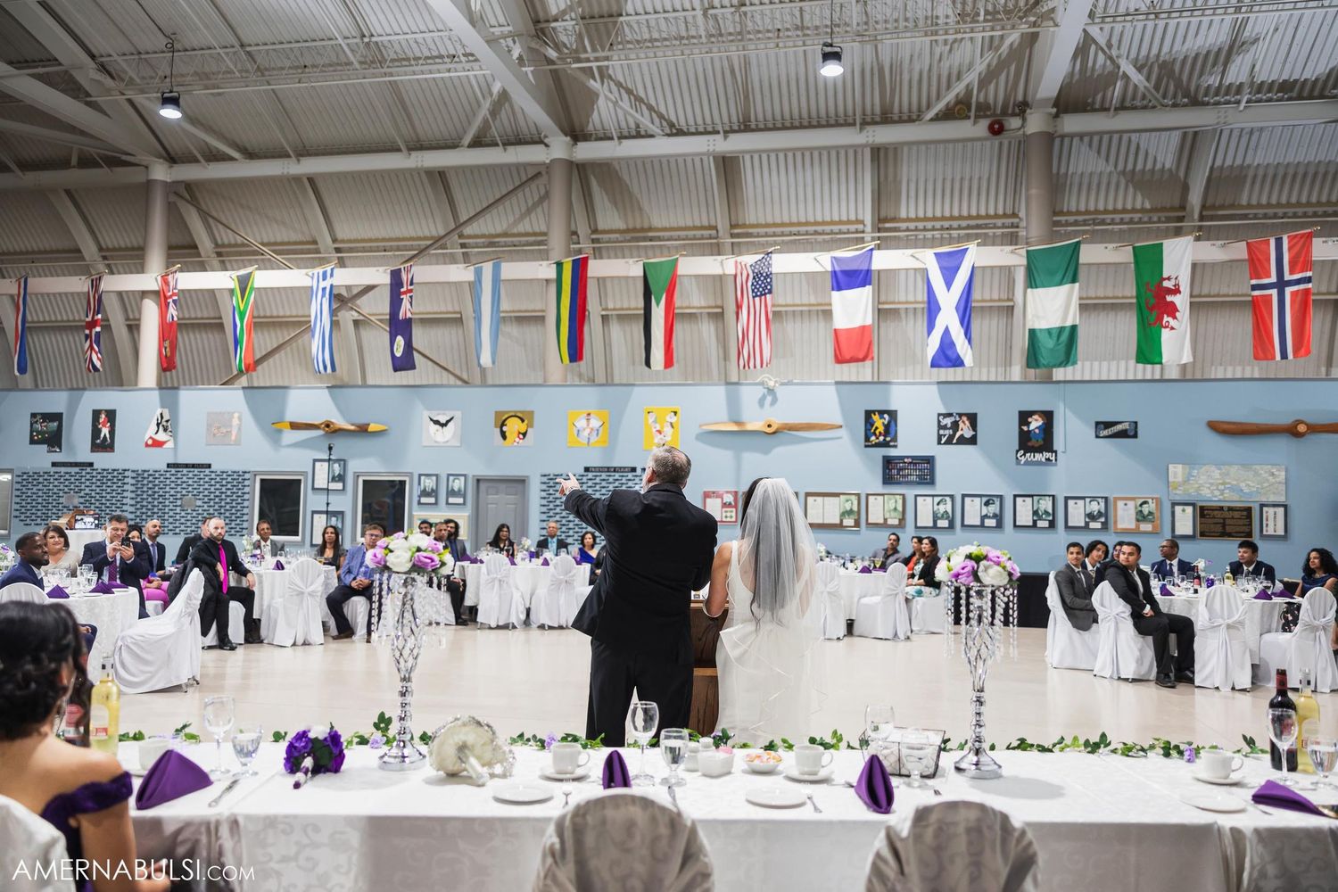 International flags hang from ceiling at elegant wedding reception in aircraft hangar.