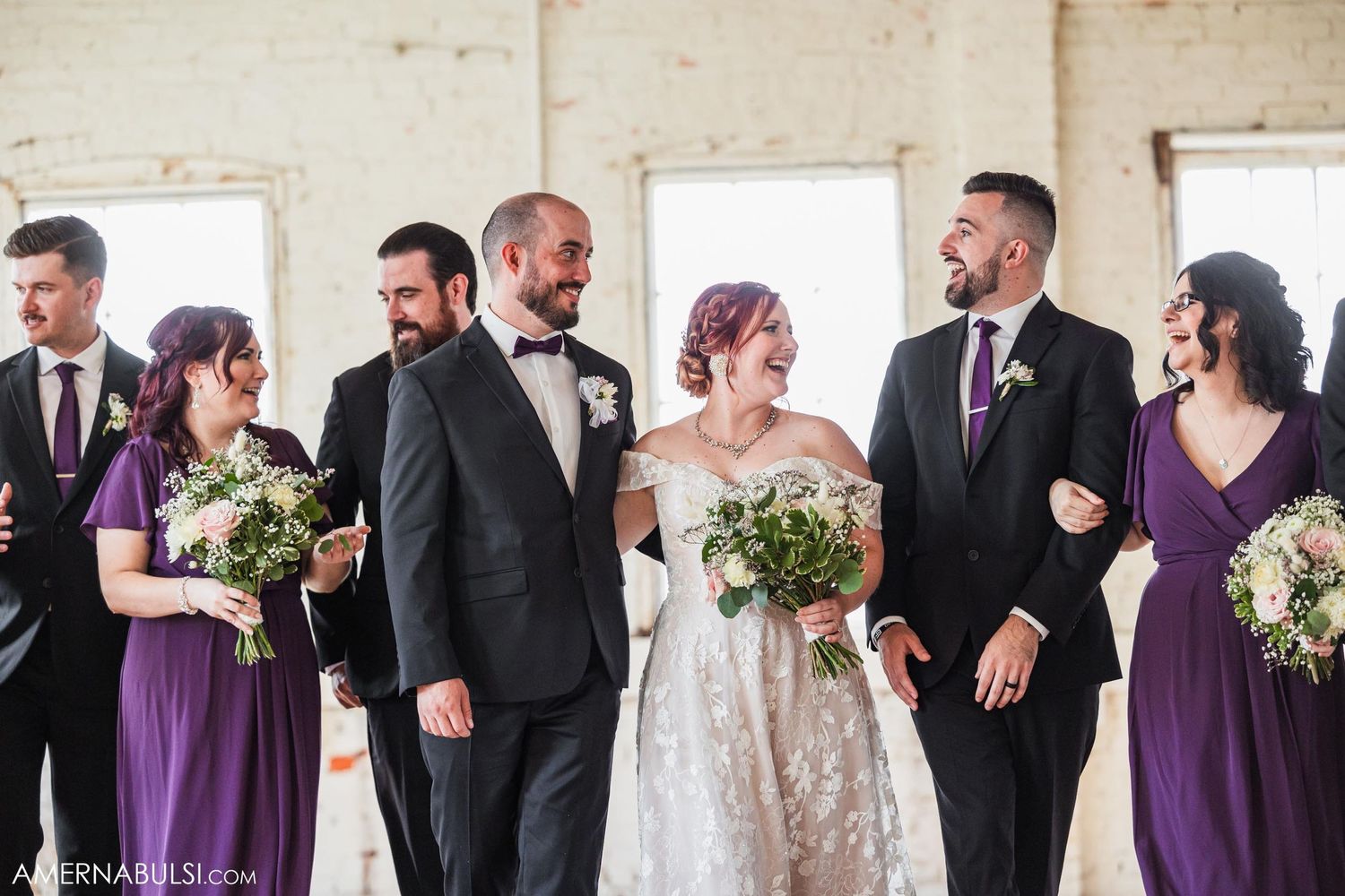 Wedding party poses casually in bright industrial space with purple and black attire.