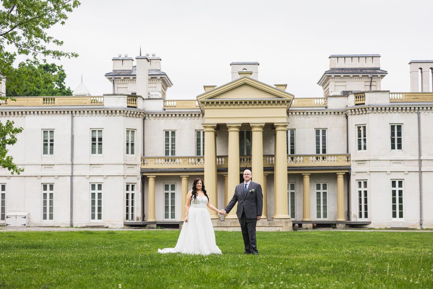 Bride and groom pose in front of a grand historic mansion with white columns and neoclassical architecture.