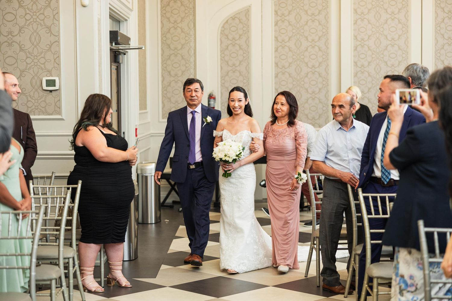 Wedding ceremony processional in elegant ballroom with checkered floor and seated guests.