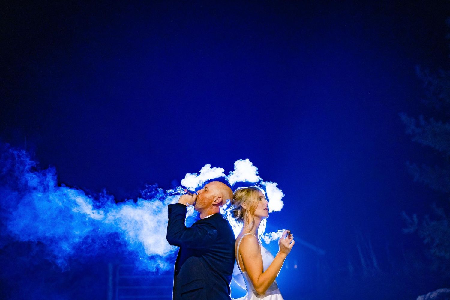 A wedding couple embraces against a dark blue sky with dramatic smoke effects.