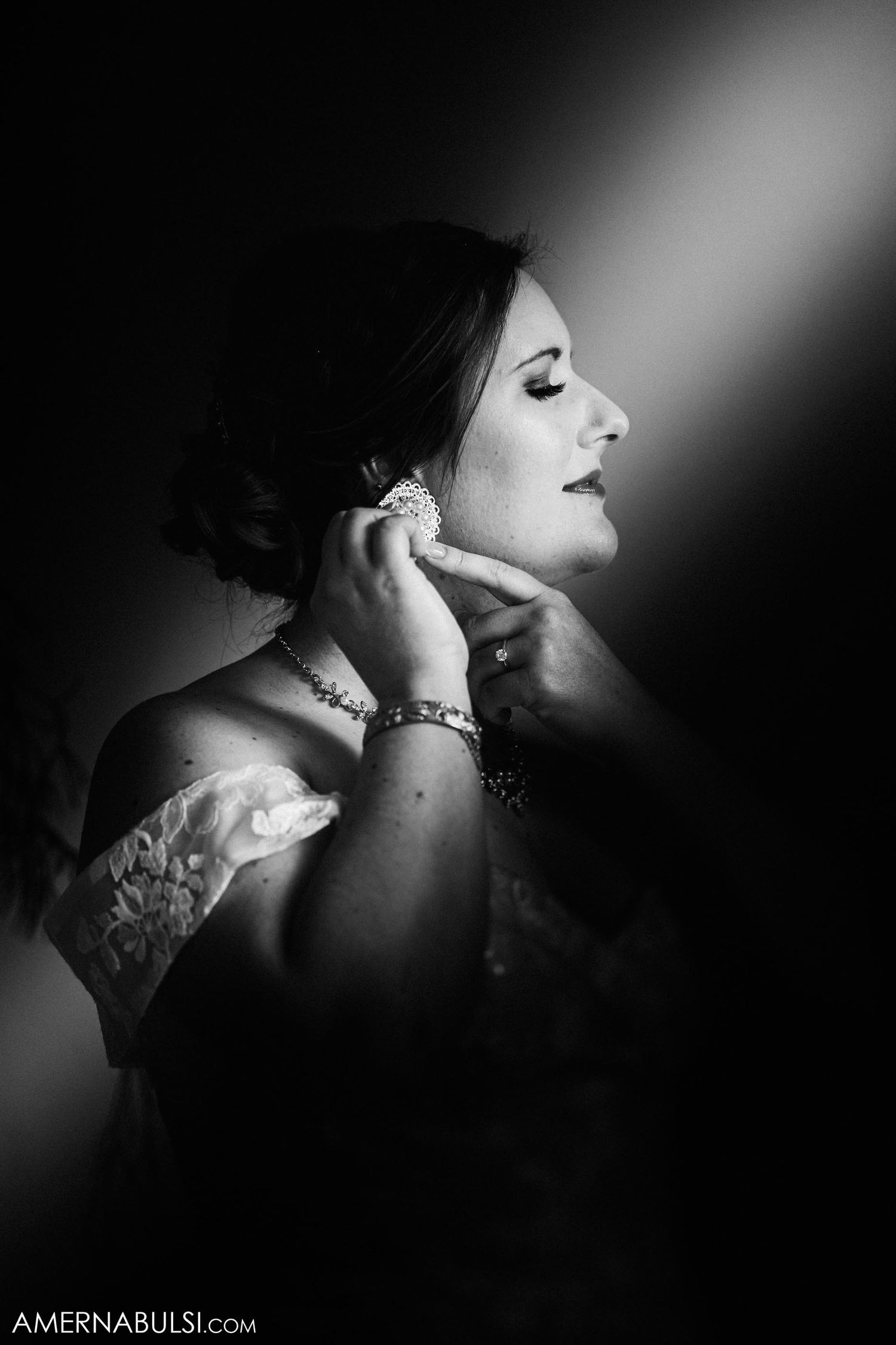 Dramatic black and white portrait of a bride putting on earrings in moody lighting.