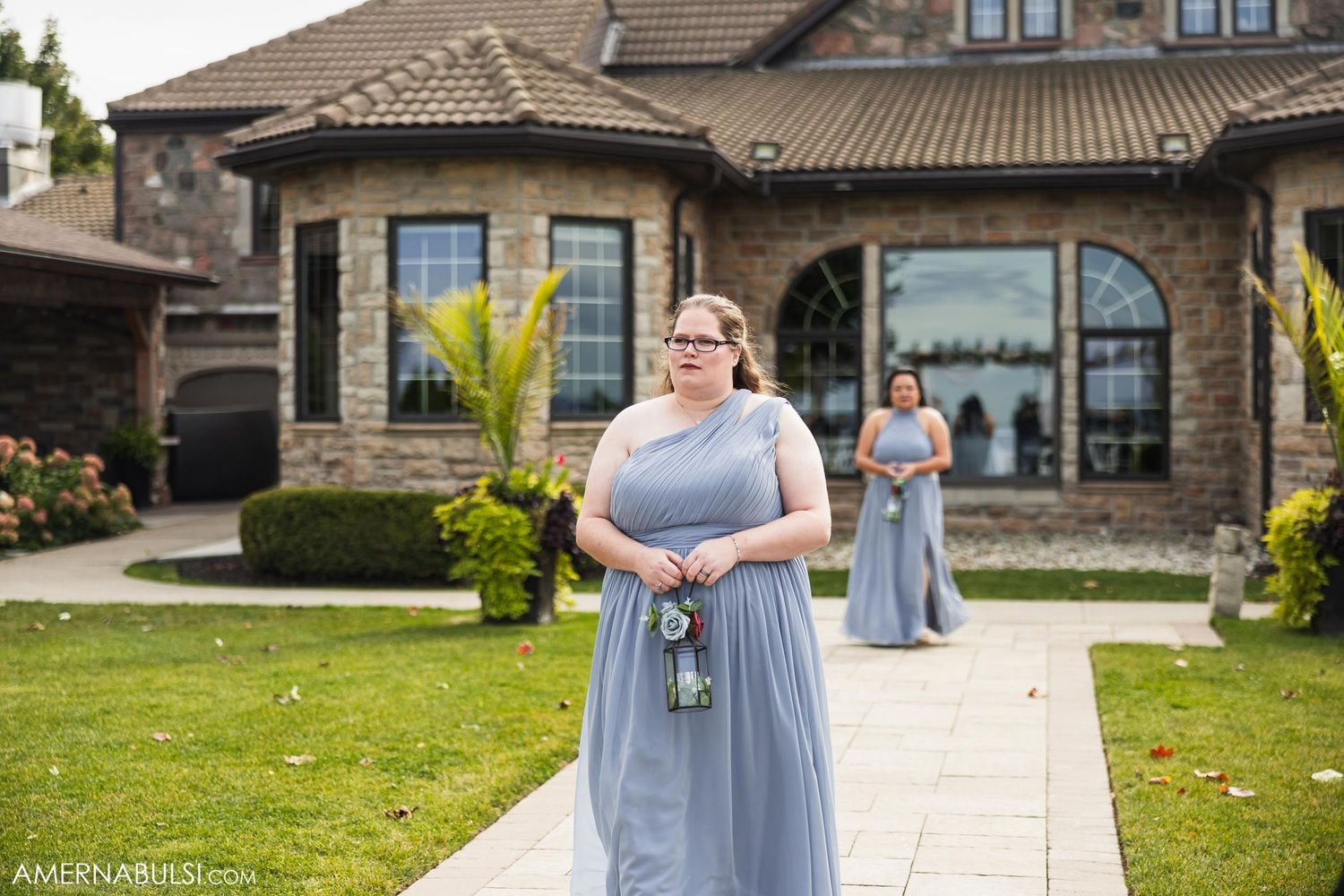 Wedding party members in light blue dresses stand outside an elegant stone house.