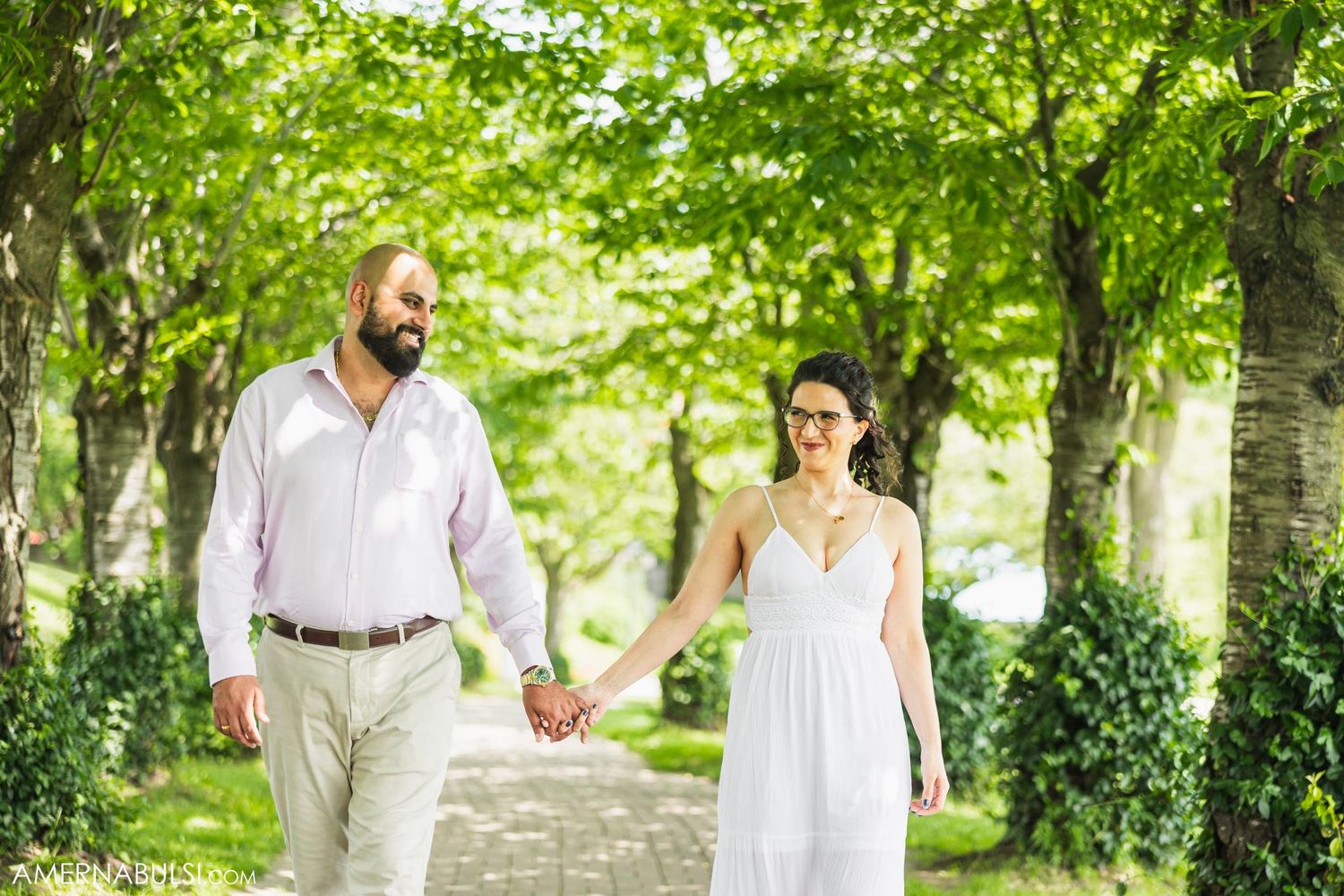 A couple in wedding attire walks hand in hand down a tree-lined path on a sunny summer day.