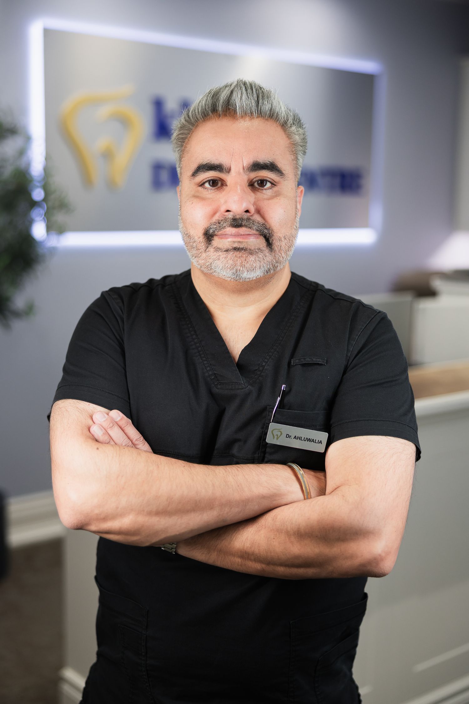 A confident dentist in black scrubs stands with arms crossed in front of a dental centre logo sign.