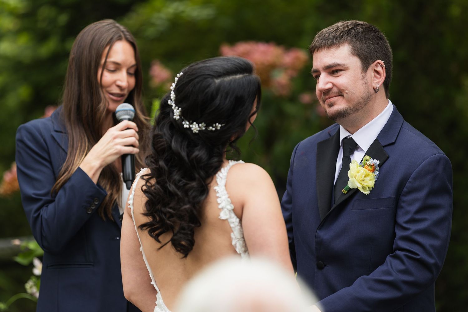 Wedding officiant speaking into microphone while bride and groom exchange vows at outdoor garden ceremony.