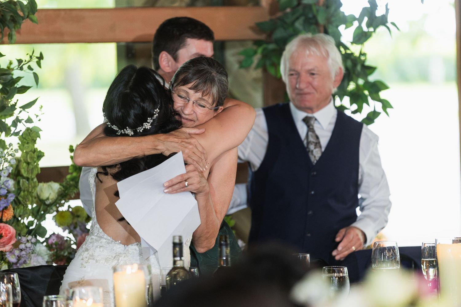 Bride and groom sharing emotional hug with guest during wedding reception speeches at rustic venue.