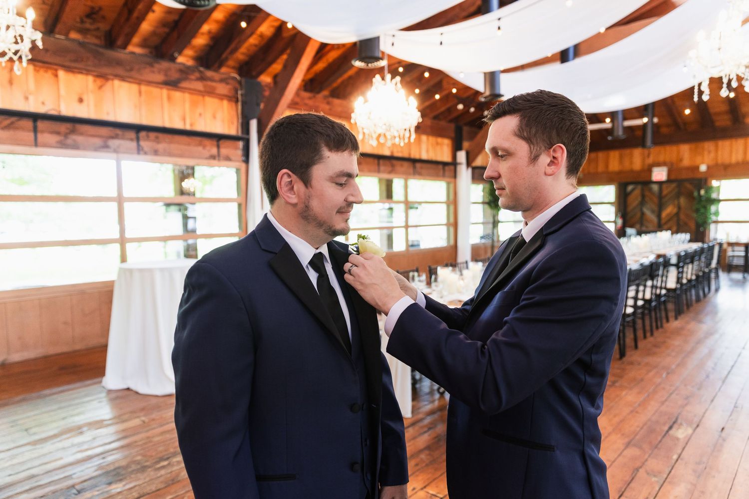 Two groomsmen in navy suits preparing for a wedding ceremony inside a rustic wooden venue with chandeliers.