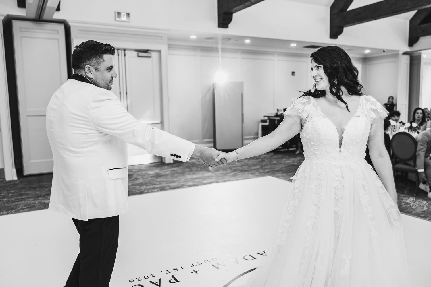 Black and white photo of bride and groom sharing first dance, holding hands at elegant wedding reception.