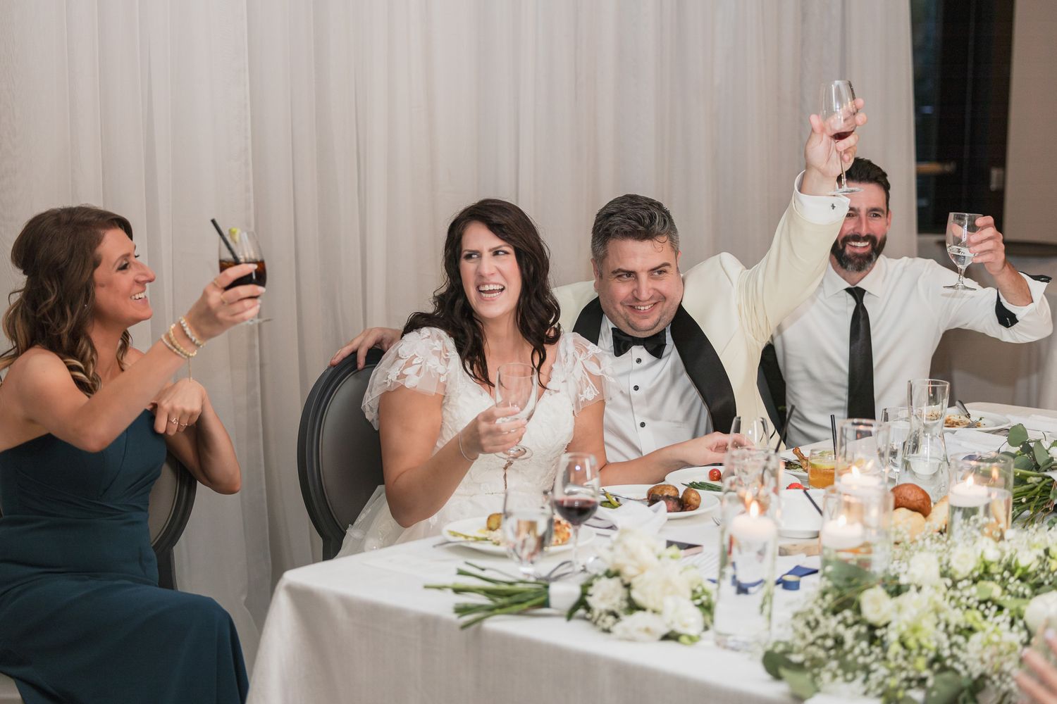 Wedding party raises glasses in a toast at the reception dinner table with candles and floral centerpieces.