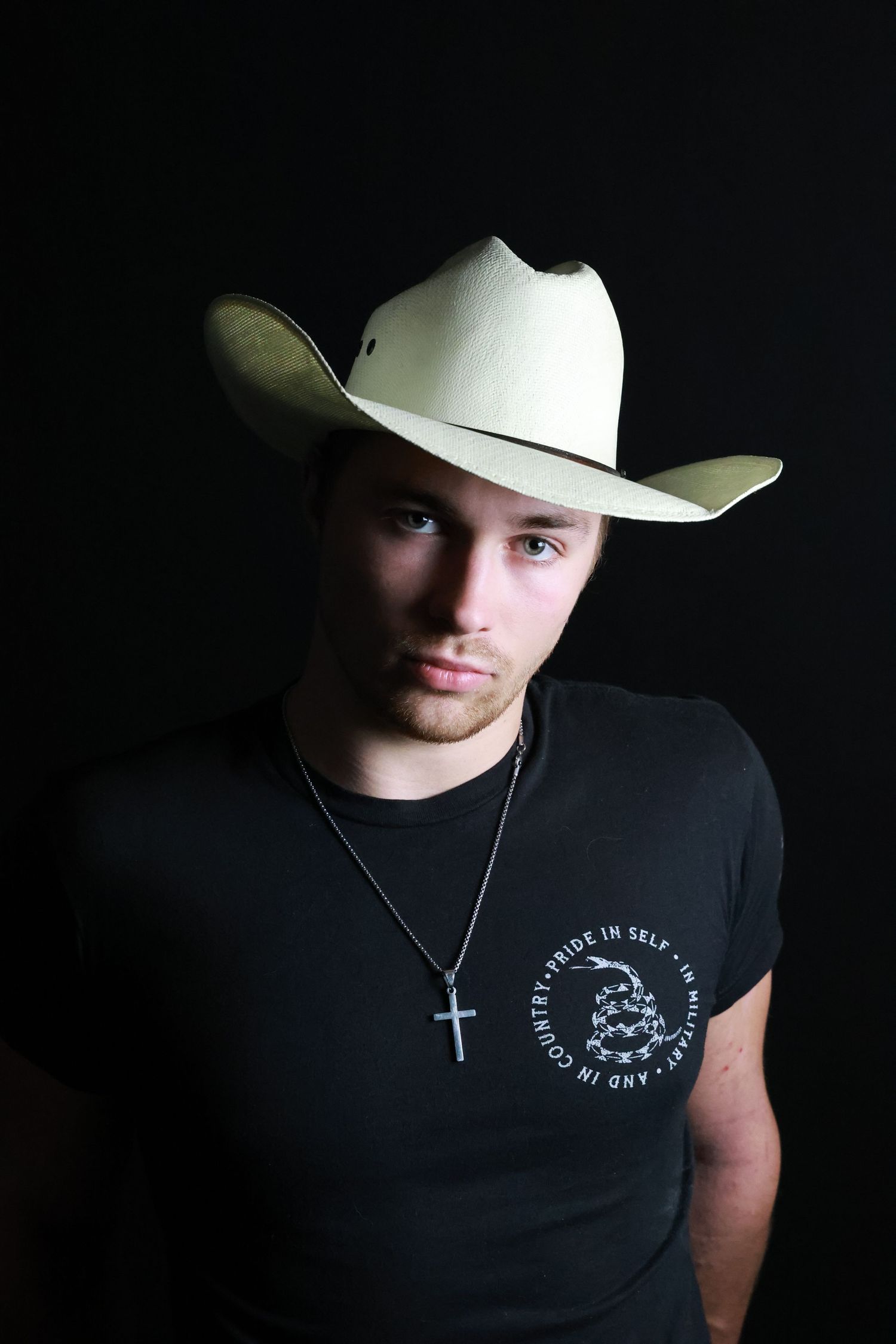 Person wearing a white cowboy hat and black sleeveless shirt with a cross necklace in a dark setting.