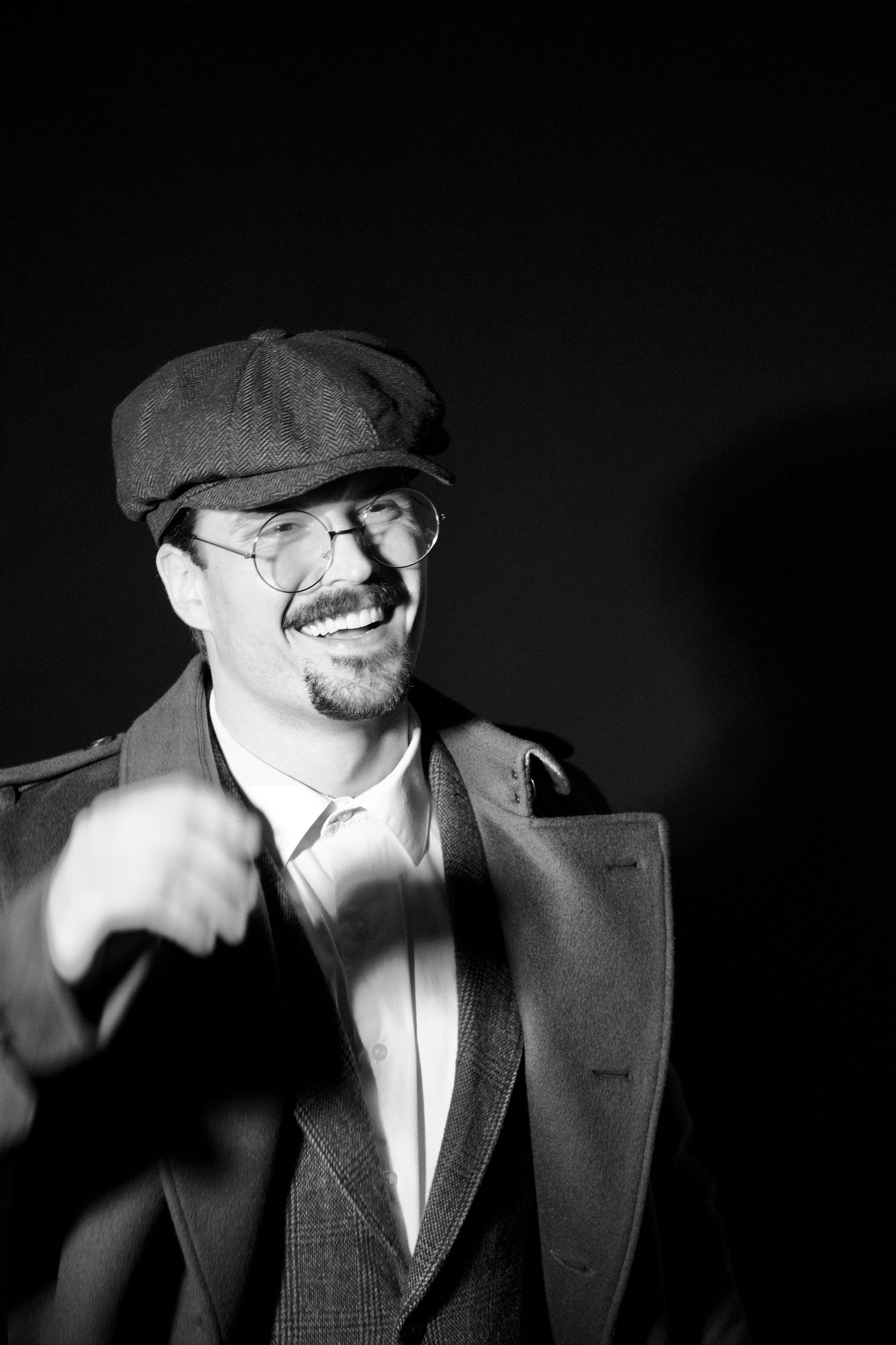 Black and white photo of a laughing man in vintage flat cap, glasses, and trench coat on dark background.