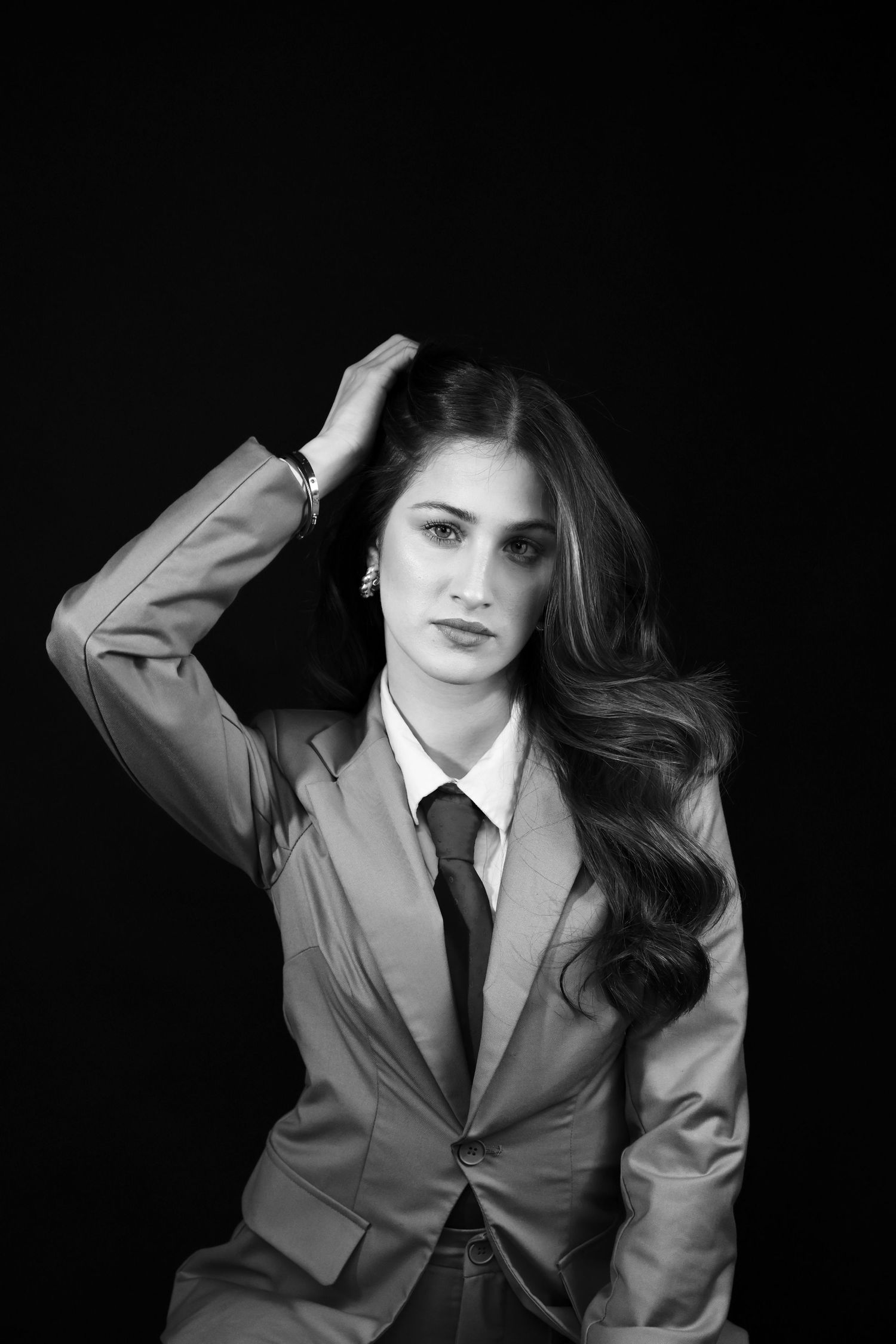 Elegant woman in blazer and tie poses with hand in hair in a dramatic black and white studio portrait.