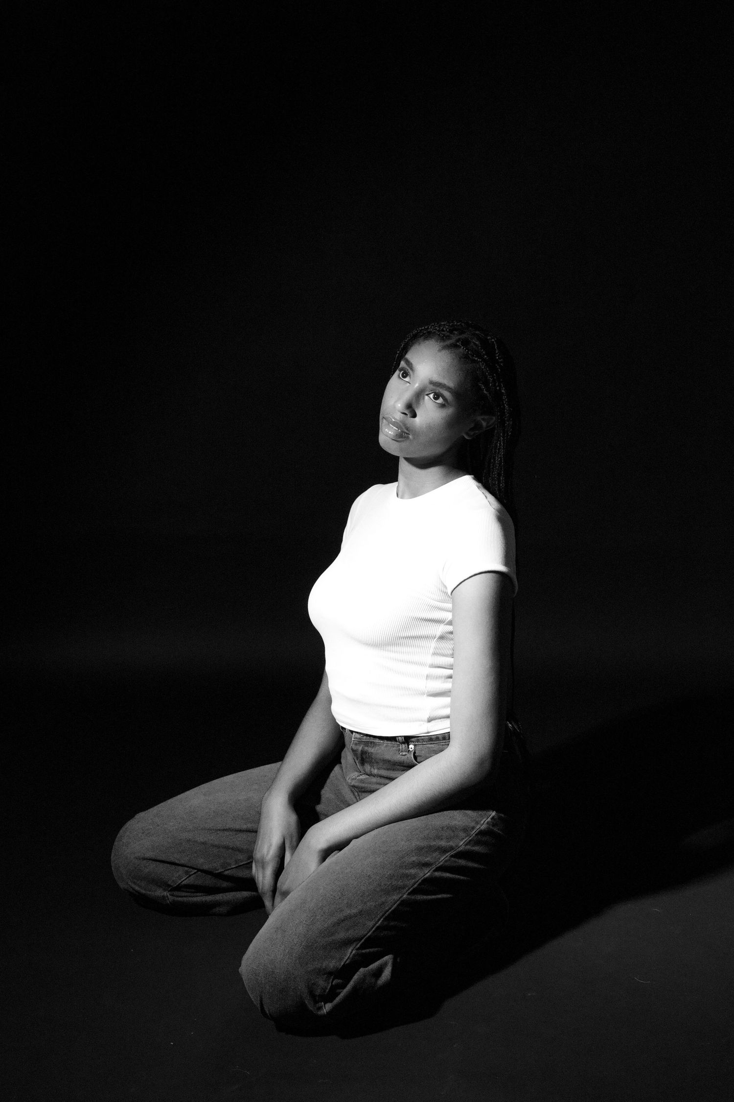 Black and white portrait of a young woman sitting on floor in white t-shirt and jeans against dark background.